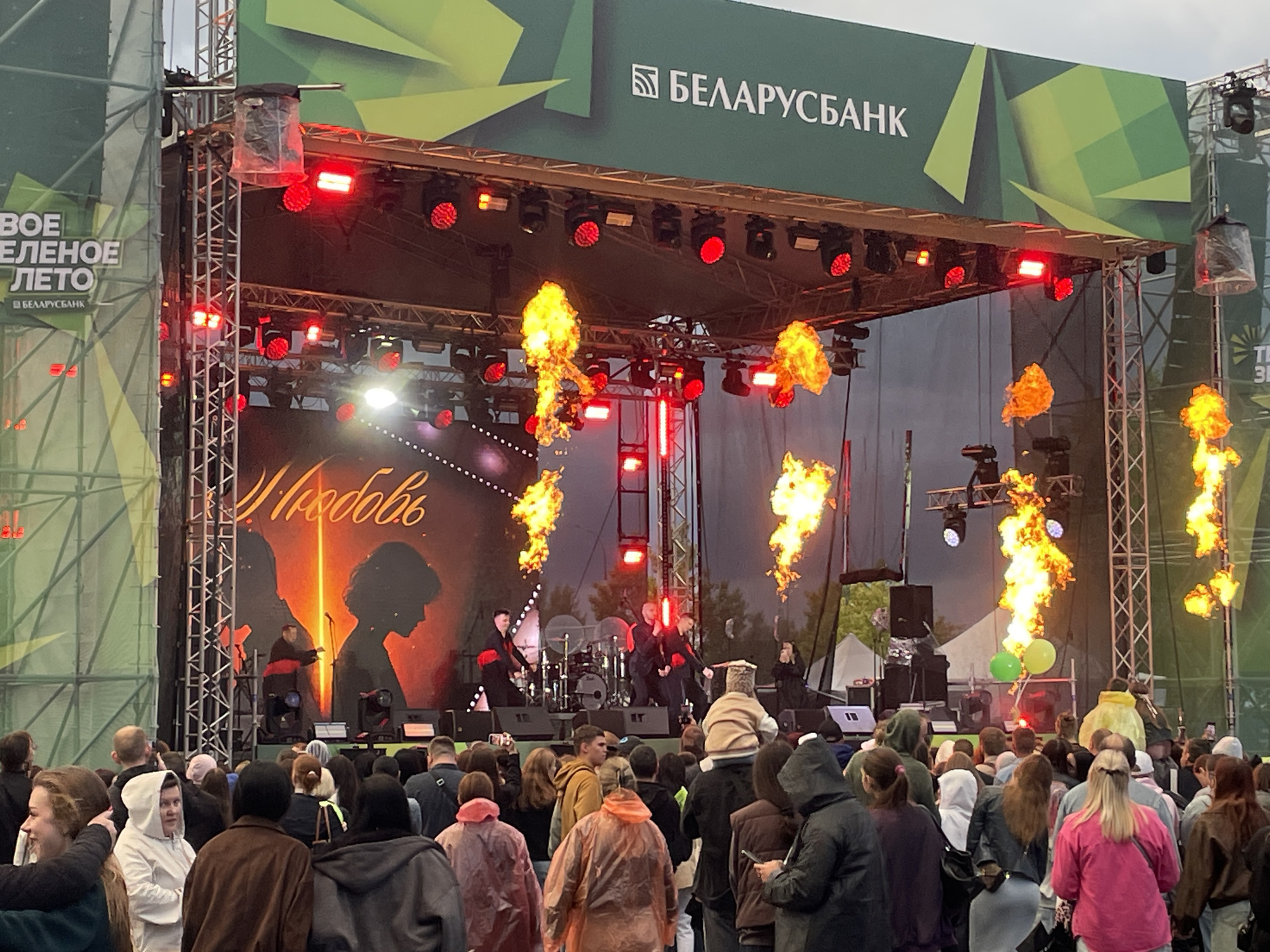 This image captures an outdoor concert event at the Brest Fortress in Brest, Belarus. The focal point is a brightly lit stage with pyrotechnic displays of fire erupting from the left and right sides. On the stage, several performers are visible, including drummers and vocalists, dressed in dark attire with red accents. The backdrop of the stage features a large graphic with the word "Любовь" (Lyubov, meaning "Love") written in elegant script, and silhouettes of a couple. Above the stage, a large green banner displays the logo and name "БЕЛАРУСБАНК" (Belarusbank). In front of the stage, a large crowd of spectators stands, many wearing casual clothing, and some with ponchos, suggesting the possibility of rain. The overall atmosphere is energetic, with the dramatic lighting and fire effects contributing to a festive and engaging concert experience.