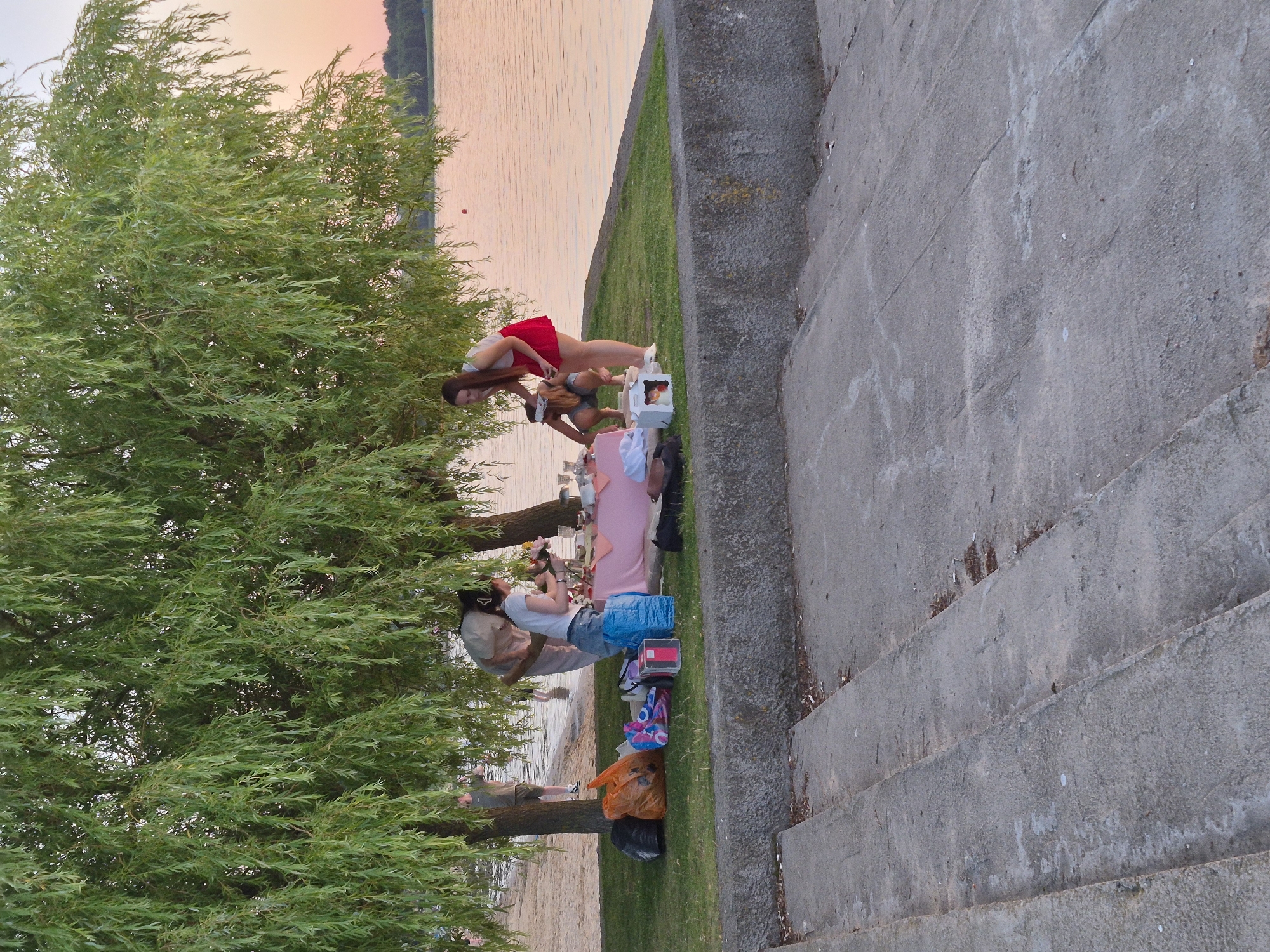 This image depicts a picnic scene by a body of water, likely a lake or river, under the shade of a large weeping willow tree. The water appears calm, reflecting the soft hues of the sunset. In the foreground, concrete stairs lead down to the grassy area where a picnic is being set up. Several individuals are gathered around a pink tablecloth spread on the grass, arranging food and drinks. Bags of varying sizes and colors, as well as what appear to be coolers and food containers, are scattered around the picnic area. In the background, a sandy beach area is visible, with people enjoying the waterfront. The overall atmosphere suggests a relaxed, end-of-day gathering. The location is identified as Minsk, Belarus.
