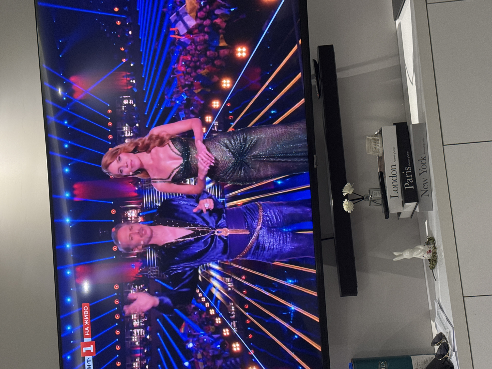 The image shows a television screen displaying a live broadcast of a show, likely a competition or performance, with a man in a blue sequined outfit and a woman in a shimmering green dress. The show features a stage with blue and gold lighting, and an audience in the background. The bottom left corner of the screen shows the text "1 HA ЖИВО," indicating it is a live broadcast on a television channel.
To the right of the TV is a white shelf with decorative items, including three books labeled "London," "Paris," and "New York." There is a small vase with artificial flowers, a small white bunny figurine, and a pair of sunglasses.
The image's location is unknown; it doesn't provide any information to infer that the location of the image is in Burgas, Bulgaria.