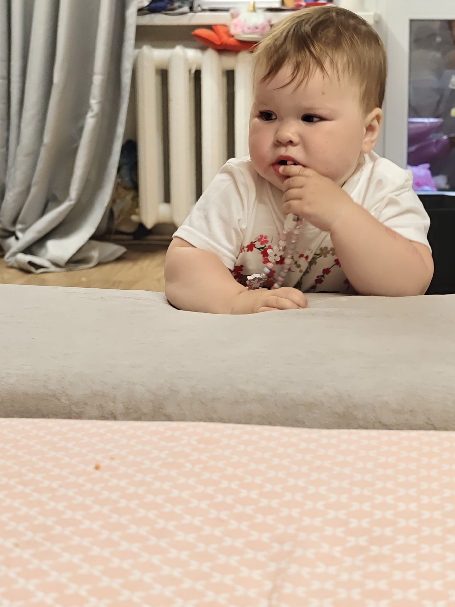 A young child is positioned behind a low, light-gray padded barrier, likely a sofa edge, in an interior setting. The child, with fair skin and light brown hair, is wearing a white shirt with a floral design and a string of beads. The child's left hand is raised to their mouth, with a finger inserted between their lips.
The background includes a radiator and curtains. A shelf with objects including toys is visible above the radiator. The foreground features a patterned surface, possibly a tablecloth.
The city of Chaikovsky, Russia is not identifiable within this image.