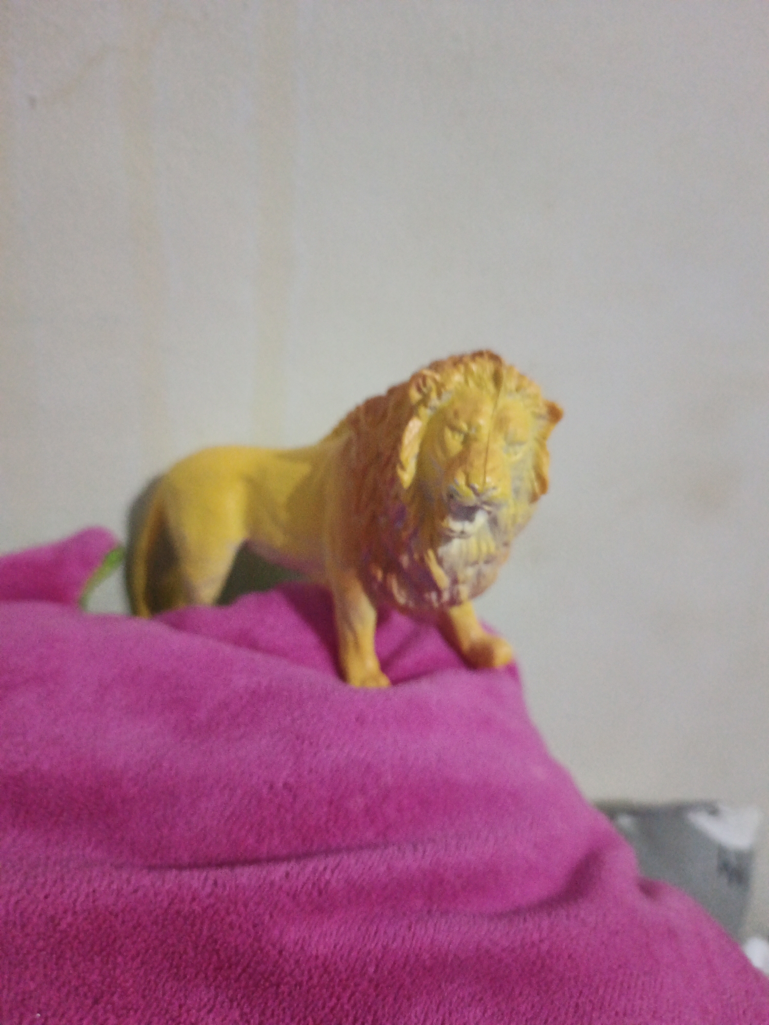 A plastic toy lion with a yellow body and a brown mane is positioned on a folded pink blanket. The lion is facing right, with its head slightly turned towards the viewer. The background consists of a plain, light-colored wall. The image appears to be taken indoors, with soft lighting. The scene is devoid of any specific location markers or identifiable elements that would place it in South Jakarta, Indonesia.