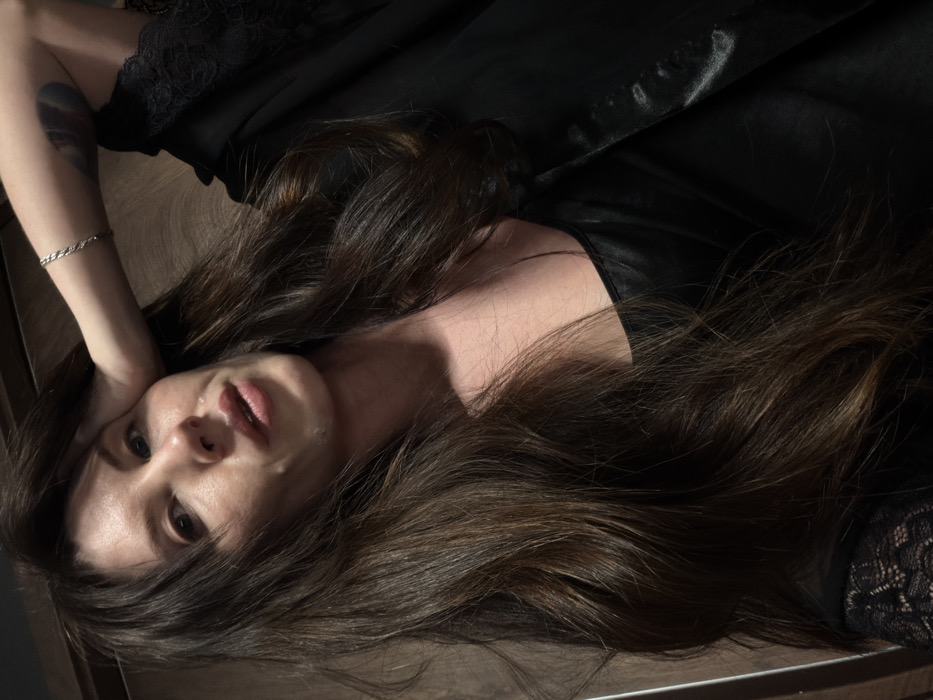 A brunette woman with fair skin reclines, her head tilted back. She's adorned in a dark garment, possibly satin, and lace embellishes the edges of her sleeves. One arm is raised, revealing a bracelet and a tattoo. Her hair is tousled and spread around her. Her expression appears solemn, with slightly parted lips. The background features a wooden floor and dark fabric.