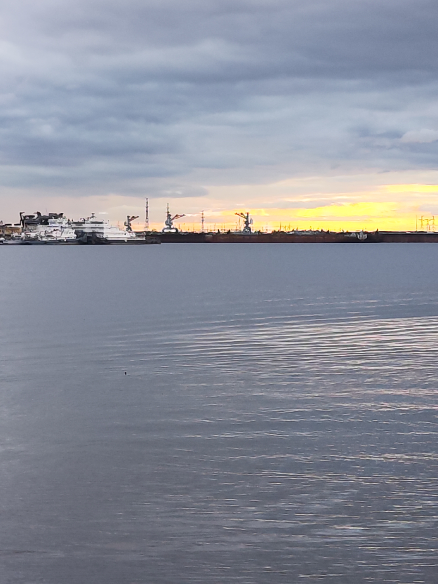 This image depicts a harbor scene at dusk, likely in Chaikovsky, Russia. The foreground is dominated by a large expanse of calm, rippling water, reflecting the muted colors of the sky. In the midground, a collection of large ships and industrial structures are visible along the waterfront. Several cranes, typical of a port, are prominent among the vessels. The largest vessel, a white ship, appears to be a ferry or a passenger liner, with multiple decks and structures. Behind it, other ships and docks can be seen, hinting at a busy maritime hub.
The sky overhead is dramatic, a mix of dark, overcast clouds and a vibrant band of yellow and orange light near the horizon, indicating the setting sun. This contrast creates a moody and atmospheric ambiance. The time of day is clearly evening, with the fading light casting long shadows and bathing the scene in a warm glow.
The overall impression is one of industrial activity at the end of the day. There is a sense of quietude and transition as daylight gives way to night. No people are clearly discernible in the image, but the presence of ships and cranes suggests human activity. The location cues, such as the type of industrial infrastructure and the natural light, are consistent with a port city like Chaikovsky. No visible text is present in the image.
