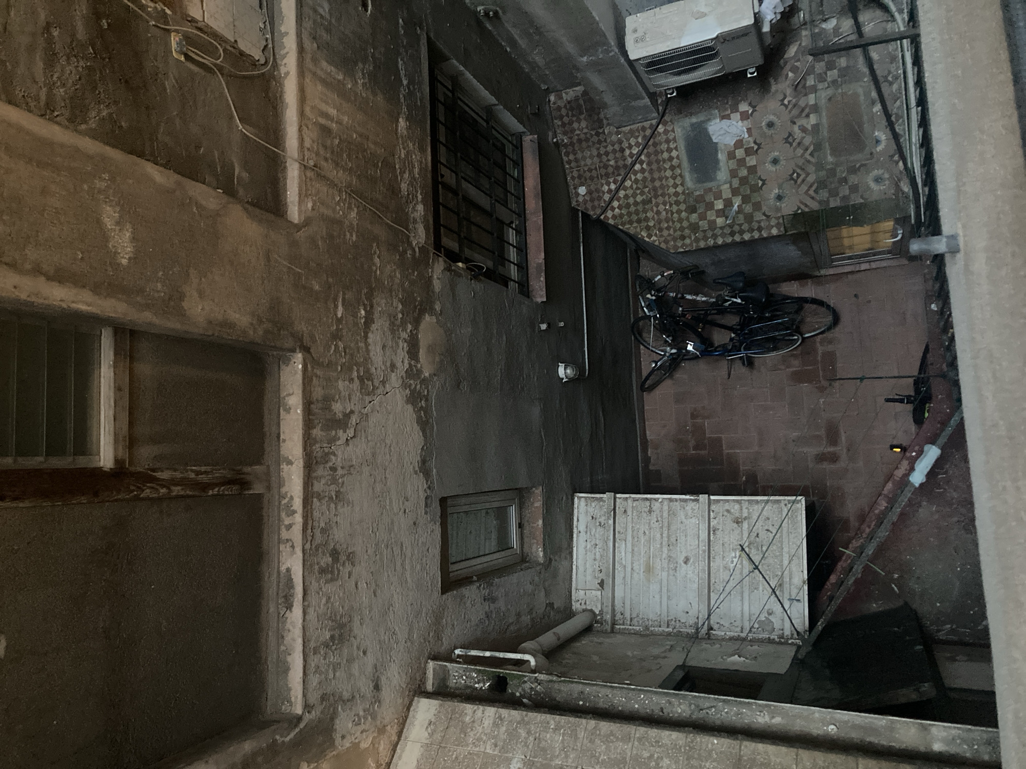 This is an exterior, overhead, high-angle shot of a grimy, aged urban courtyard or alleyway in Barcelona, Spain. The scene is dominated by deteriorating concrete structures, with cracked and peeling surfaces indicative of disrepair. Light filters in from above, illuminating the space dimly.
In the foreground, a light-colored concrete ledge runs along the bottom of the frame. To the left, a section of a grimy, tiled wall is visible, with a pipe running vertically along it. Below this, a white, slatted cabinet or structure is mounted on the wall, appearing weathered and dirty, with wires attached to it.
Further into the courtyard, the floor is covered in a mix of red brick tiles and patterned square tiles in shades of brown and white. Several bicycles are leaning against the wall on the left, with their wheels visible. Some bags are attached to the handlebars.
In the center and right of the frame, more of the courtyard floor is visible, including more patterned tiles and some embedded rectangular shapes, possibly grates or access panels. A metal grate is affixed to an opening in the ceiling. An air conditioning unit is mounted on the wall on the far right.
The overall impression is one of neglect and a forgotten urban space. The lighting is muted, with harsh shadows and areas of darkness. No people are visible in the scene.