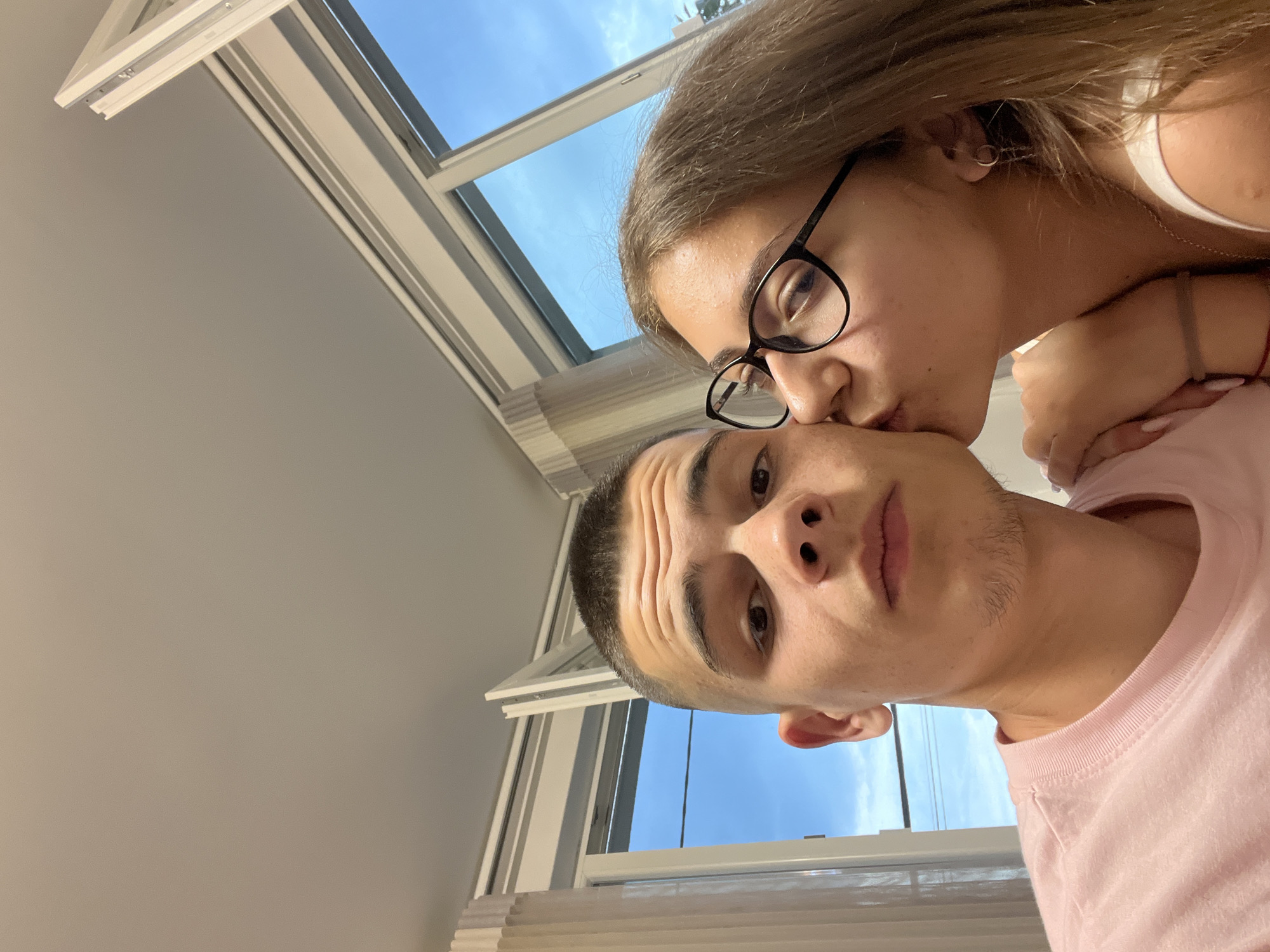 The image captures a close-up selfie of two young individuals in an indoor setting in Karavelovo, Bulgaria. On the left, a male with a short buzz cut, prominent forehead wrinkles, and faint facial stubble wears a light pink crew-neck t-shirt, looking directly into the camera. To his right, a female with long light brown hair and black-rimmed eyeglasses is kissing his right cheek. She wears a white tank top, multiple earrings in her right ear (a hoop and a dangle), and has a thin red string bracelet on her left wrist. Her left hand, with light-colored nail polish, rests on the male's upper left arm. Behind them, a large window with white frames reveals a clear blue sky with scattered white clouds, suggesting daytime. A thin dark line, possibly a utility wire, traverses the sky horizontally through the window. White, vertically pleated curtains are visible on the left side of the window frame. The ceiling is plain and light-colored.
