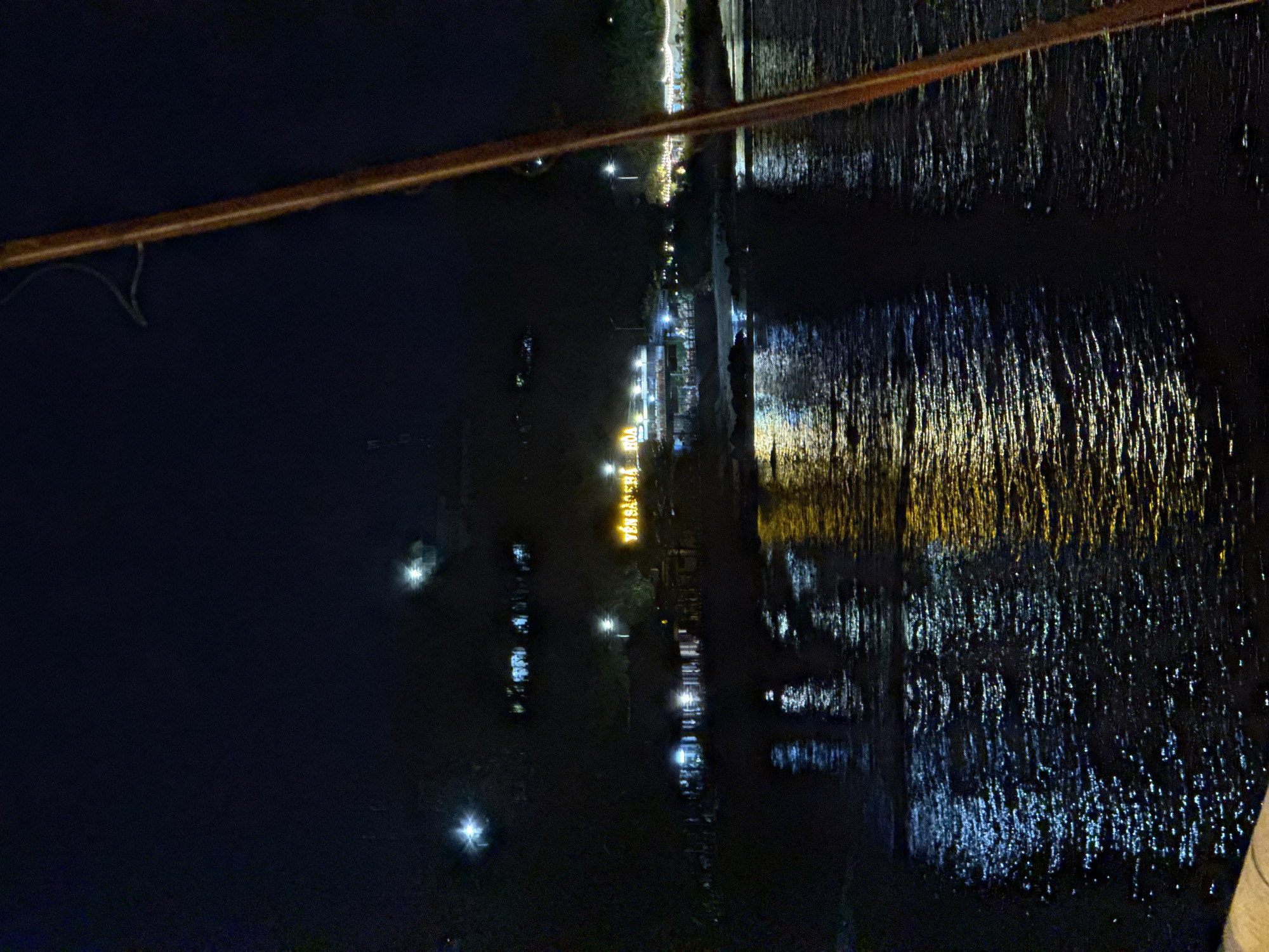The image depicts a nighttime view of a waterfront area in Nha Trang, Vietnam, likely along the Trần Phú beach promenade. On the right side of the frame, a thick, reddish-brown wooden pole, possibly part of a pier or a boat, runs vertically, partially obscuring the scene. The background is dark, with the lights from buildings and street lamps creating reflections on the water in the foreground.
A building complex with illuminated windows and signs is visible across the water. One sign prominently displays yellow text that reads "YEN SA CHA HOA". This suggests a commercial establishment, possibly a restaurant or shop, catering to tourists. The lights from these buildings are reflected in the rippling water, creating streaks of blue and yellow. The water itself appears dark, with the reflections breaking it into a textured surface. The overall atmosphere is one of a lively urban waterfront at night.