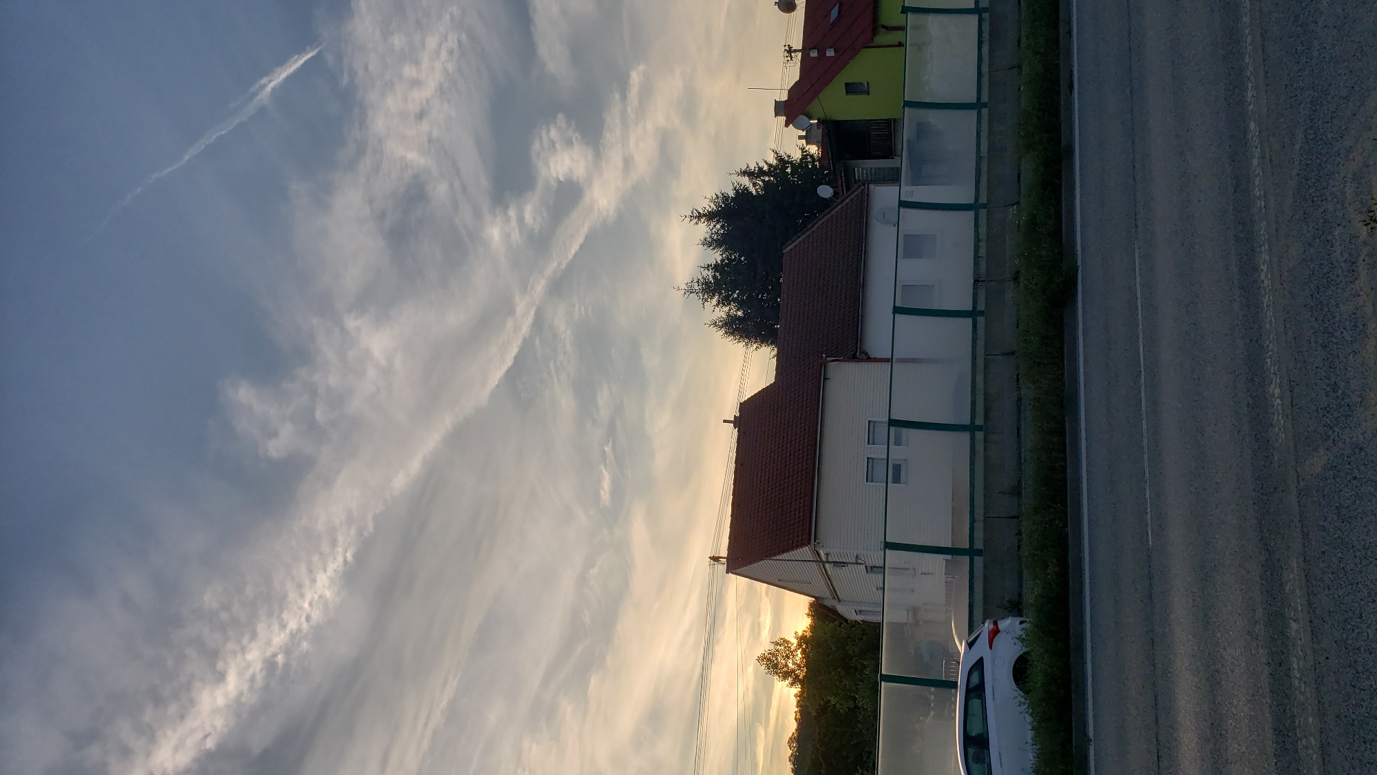 The image depicts a suburban residential scene at dusk, likely on the outskirts of Prešov, Slovakia. The sky is filled with streaky clouds and a faint contrail, suggesting either sunset or sunrise. In the foreground, a grey asphalt road is visible, with a white car parked on the right side of the road. Behind the road, there is a row of houses with red-tiled roofs, and a translucent green fence. The houses appear to be single or two-story dwellings. The environment is quiet and appears to be a residential area. There are no people visible in the image. The overall impression is one of peaceful, suburban life at the end of the day.