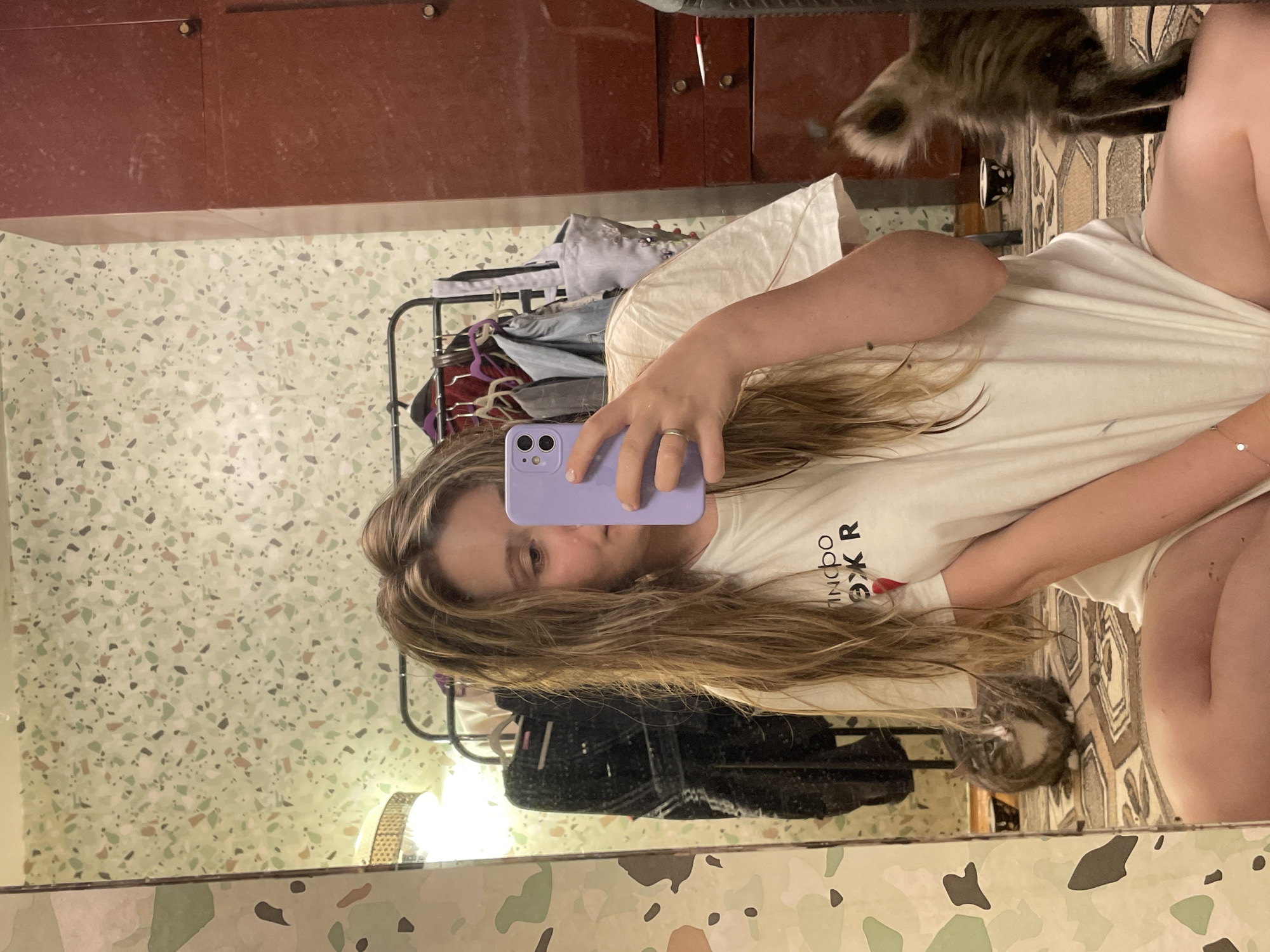 A young woman with long, blonde, wavy hair is taking a mirror selfie in a room. She is wearing an oversized white t-shirt. She is holding a lavender phone in her right hand, pointing it towards the mirror. A silver ring is visible on her left ring finger. To her left, a grey and white cat is sitting on the floor, looking towards the camera. To her right, a brown and grey cat's tail and hindquarters are visible as it walks away from the camera. The background consists of a patterned wallpaper, a clothing rack with several garments, a tall, dark red cabinet with drawers, and a floor with a patterned rug. The overall lighting is dim.