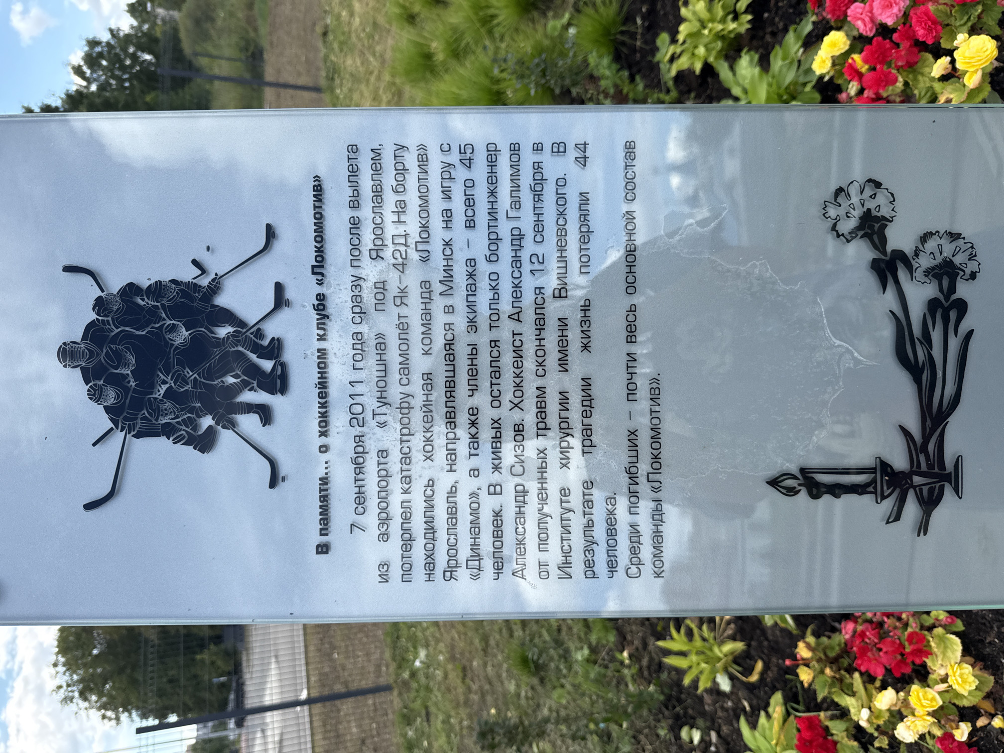 The image shows a memorial plaque with an etching of hockey players at the top, a lit candle, and two stylized flowers at the bottom. The text on the plaque, in Russian, commemorates the Lokomotiv Yaroslavl ice hockey team. It states that on September 7, 2011, the team's Yak-42D aircraft crashed near Tunoshna Airport in Yaroslavl shortly after takeoff. The text details that the team was en route to Minsk for a game against Dinamo. Of the 45 people on board, only the flight engineer survived. The text also mentions that hockey player Alexander Galimov died from his injuries on September 12 at the Vishnevsky Institute of Surgery, bringing the total loss of life to 44 people. It concludes by stating that almost the entire main roster of the Lokomotiv team was lost. The plaque is positioned outdoors, with greenery and flowers visible in the foreground and background. The surrounding environment suggests a public memorial site.