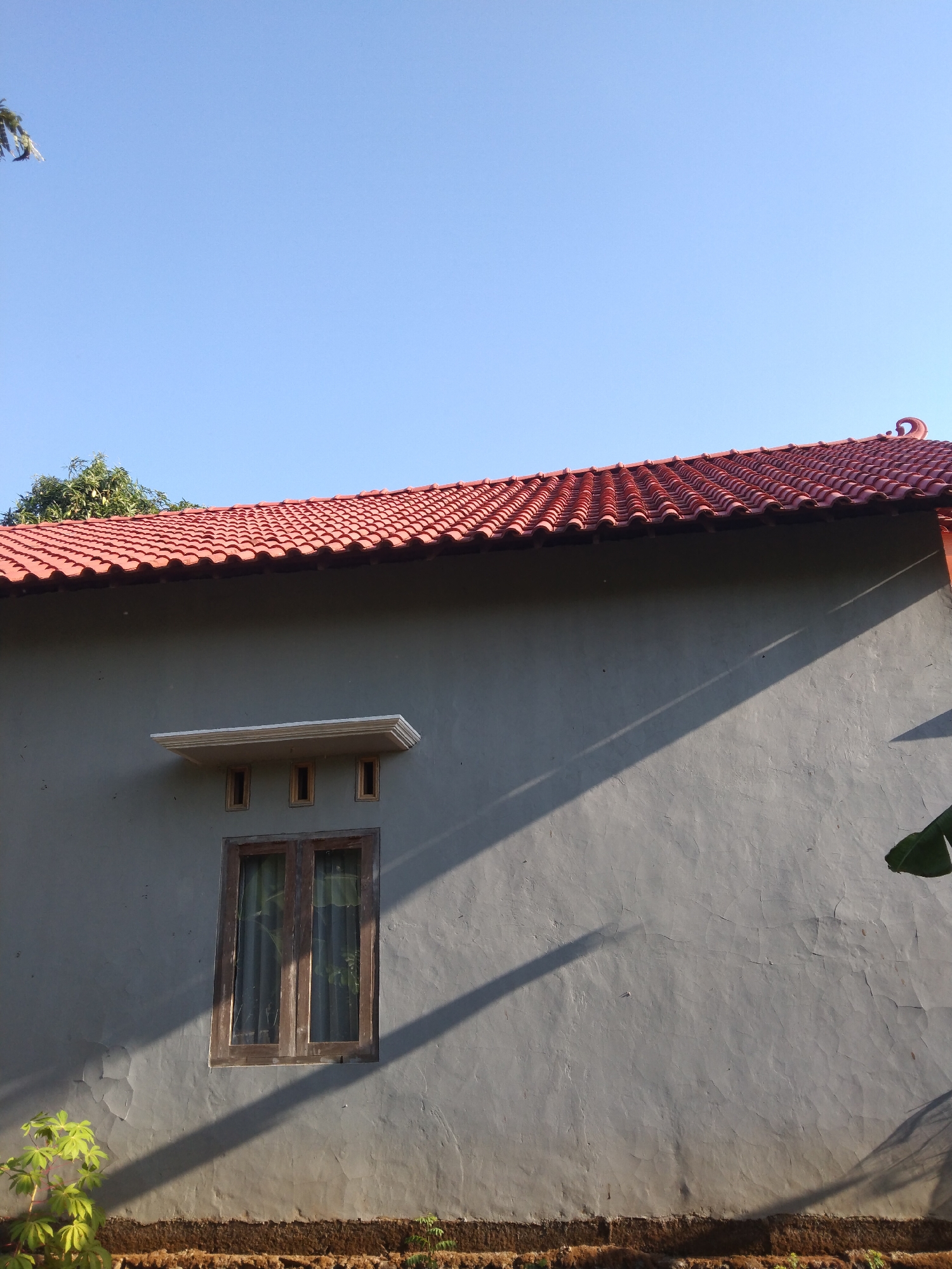 The image shows the exterior of a single-story house in Krajan, Indonesia, under a clear blue sky. The house features a red tiled roof with decorative end pieces. A single, wooden-framed window with glass panes is visible on the gray, stucco exterior wall. Above the window, there is a decorative ledge with three small, rectangular openings. A prominent shadow from an unseen object stretches diagonally across the wall from the upper right to the lower left. In the foreground, at the base of the wall, there is a strip of dark earth with some green foliage, including a small plant with large leaves on the left side and a broad green leaf extending into the frame from the right. The scene is illuminated by bright sunlight, indicating it is daytime. There are no people visible in the image.