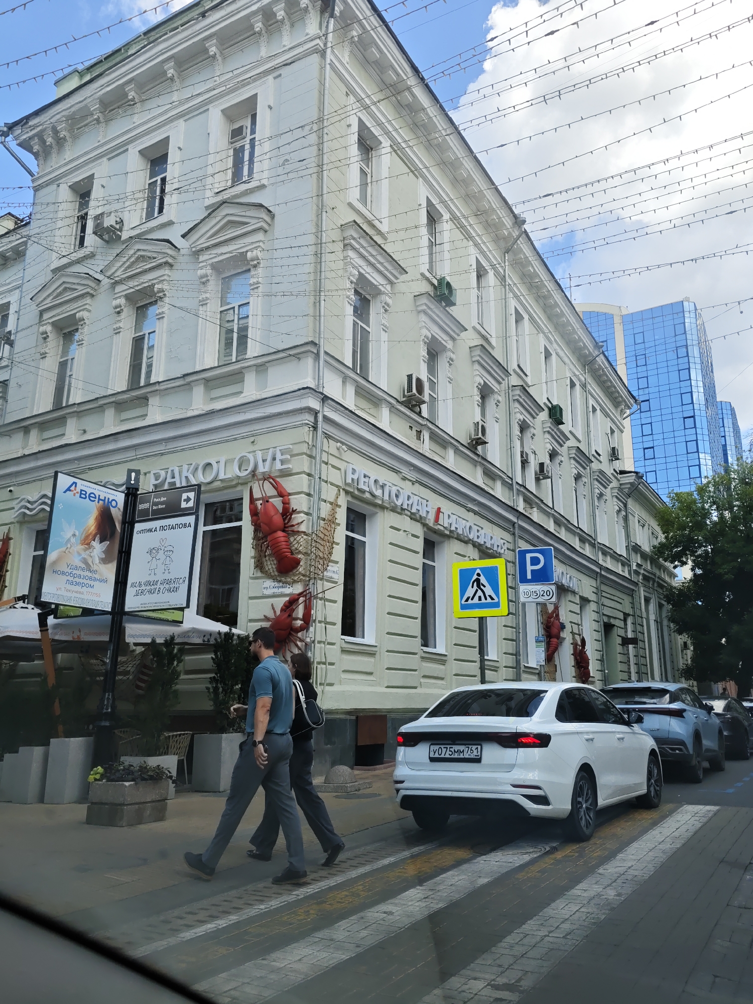 A street scene in Rostov-on-Don, Russia, features a light-colored, multi-story building with ornate architectural details, including decorative cornices and window surrounds. Strings of lights are strung across the street, possibly for decoration. A modern blue glass skyscraper is visible in the background.
On the side of the building, a sign for "PAKOLOVE RESTAURANT" is prominently displayed, along with large red lobster decorations. Several vehicles are present, including a white sedan with the license plate "У075ММ 761 RUS" in the foreground, followed by a light blue SUV and another dark vehicle.
Two individuals are crossing a pedestrian street, with the person in front wearing a blue polo shirt and grey pants, and the person behind wearing dark clothing. A pedestrian crossing sign and a parking sign are visible on a pole. The street surface is paved with cobblestones and painted markings for the pedestrian crossing. Planters with greenery are located on the sidewalk. The sky is cloudy.