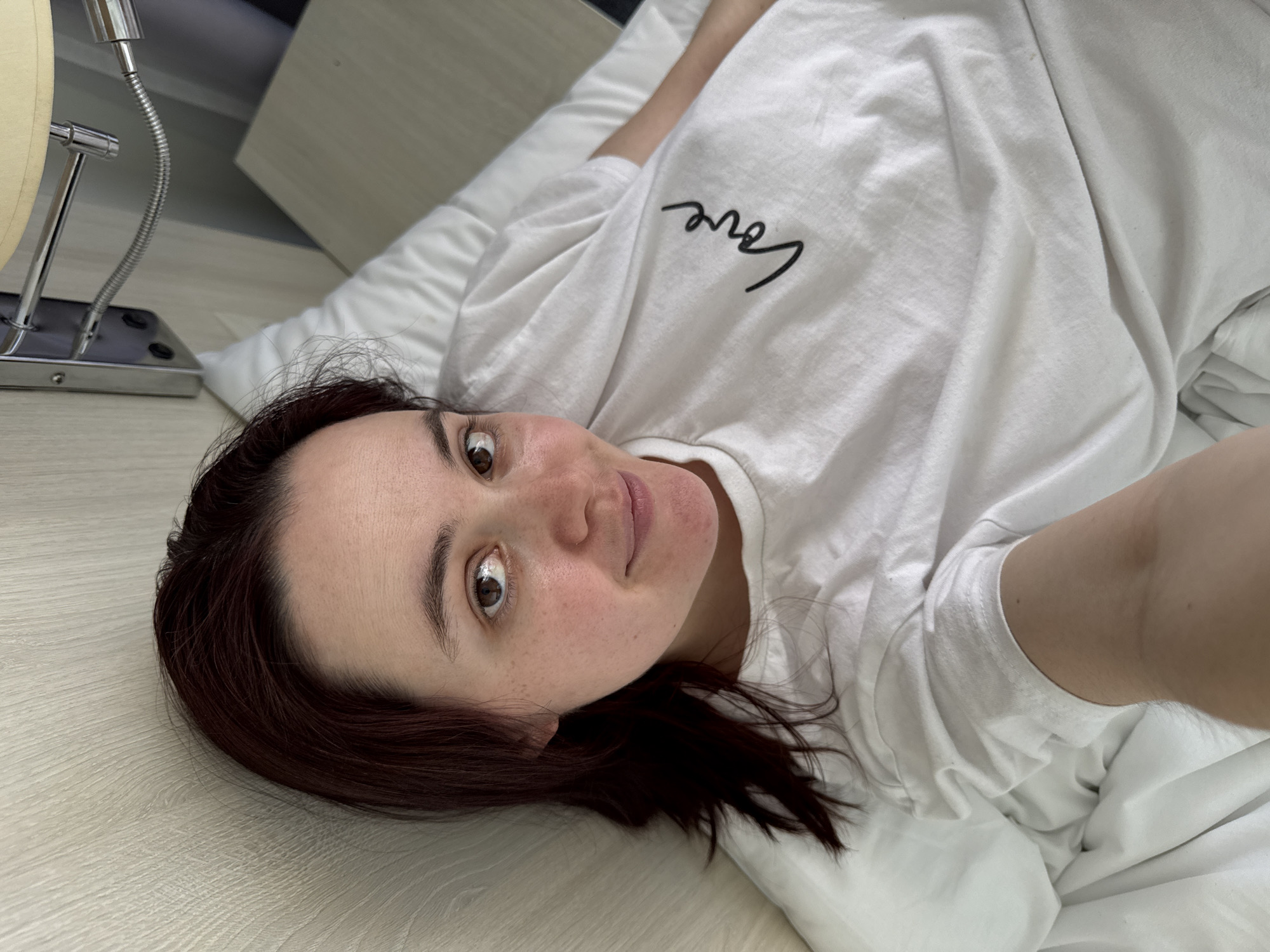 A woman with brown hair is lying on a white bed in a room. She is wearing a white t-shirt with the word "love" written on it. Her head is resting on a light-colored surface that might be a bedside table or the floor. A lamp is visible in the upper left corner of the image. The location is unknown, and there is no indication that the image was taken in Sochi, Russia.