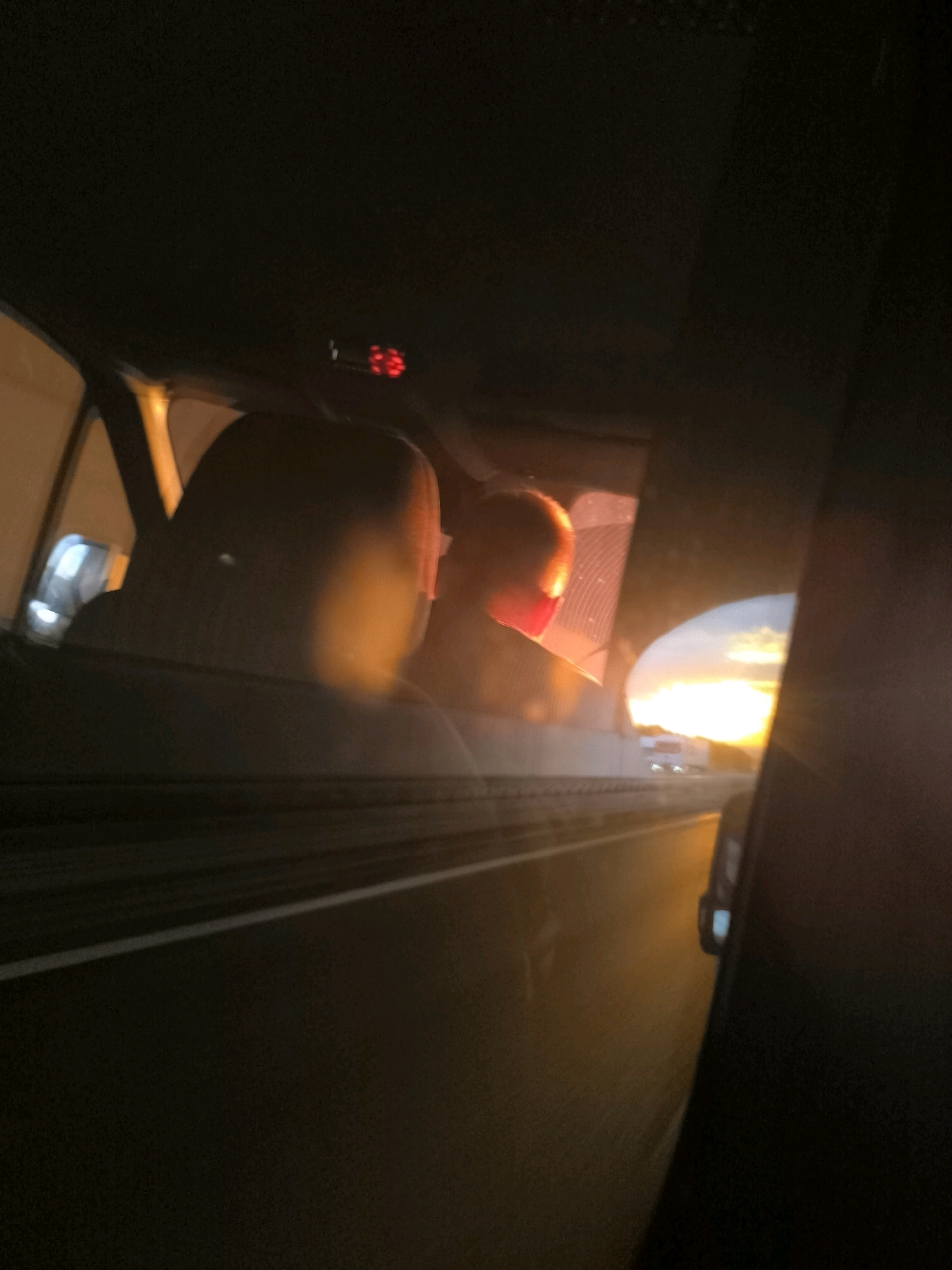 The image shows the interior of a car traveling on a highway at sunset. The perspective is from the back seat, looking forward and out the side window. Two figures, likely children due to their size, are visible in silhouette against the bright sunset. One figure, in the center of the frame, is illuminated by the sunset's orange glow, with their head tilted forward. To the left, another figure's head is partially visible. The car's interior is dark, with the dashboard and ceiling visible in the upper part of the frame. A digital clock displays red numbers on the ceiling. The highway stretches ahead, with a white line marking the lane. In the distance, a truck is visible on the highway. The sunset is casting a warm, orange light across the scene. The location is identified as a highway in the vicinity of Stawki, Poland.