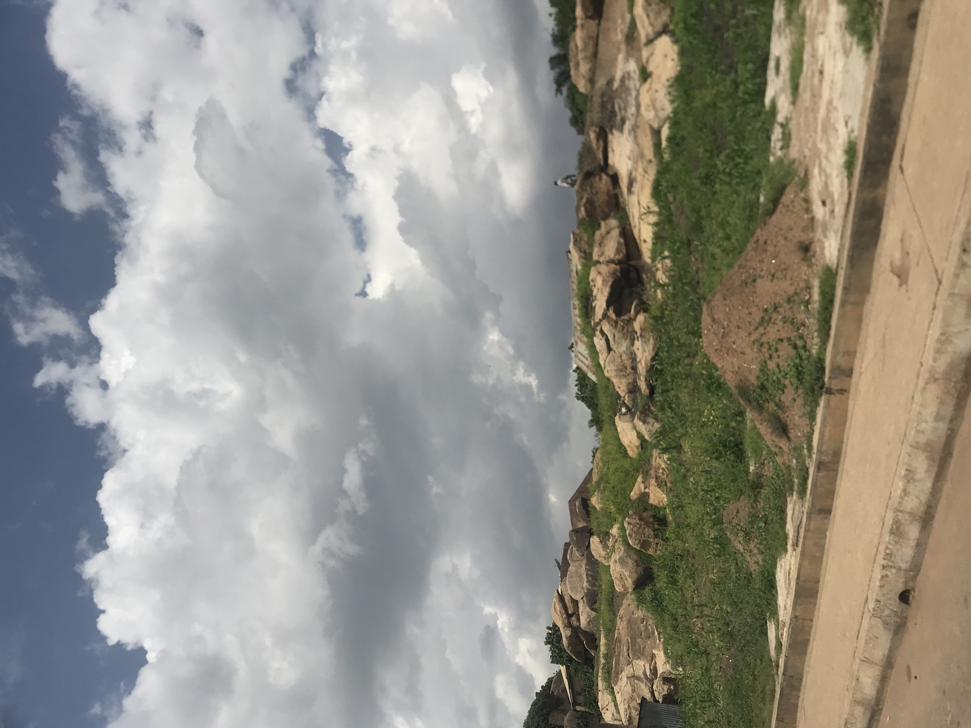An eye-level, wide shot captures a natural landscape with human elements in New Stadium, Nigeria. The scene is dominated by a cloudy sky featuring large, white cumulus clouds against a backdrop of dark gray storm clouds. Below, a rocky, hilly terrain is covered with lush green grass. Large, beige rocks are scattered across the landscape, with some forming a rough pile in the mid-ground. A small, brown dirt mound is visible in the foreground. A person, seen from behind and appearing to be on foot, stands atop one of the rock formations in the mid-ground, looking out over the scene. In the far background, hints of structures with corrugated metal roofs emerge from behind the vegetation and rocks. The foreground features a paved road with a concrete curb and drainage channel, suggesting the edge of a settlement.