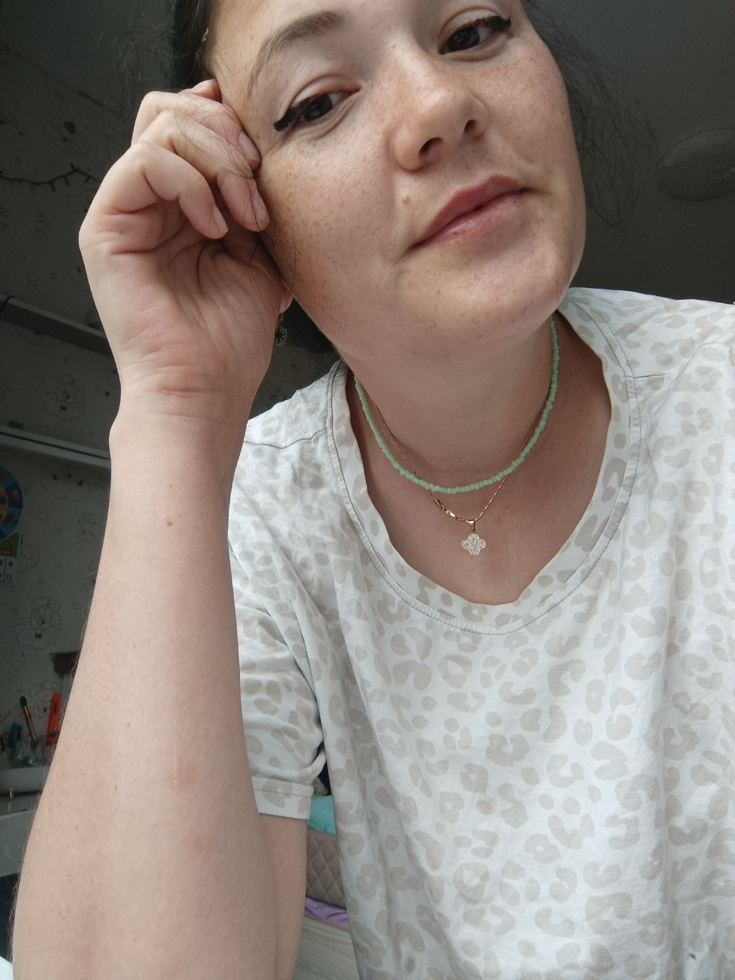 The image is a close-up, selfie-style photograph of a woman's face and upper torso. She has dark hair and is looking towards the camera with her left hand resting on her face, near her eye. She is wearing a white t-shirt with a light brown leopard print pattern. Around her neck, she is wearing two delicate necklaces, one with small green beads and another with a rose gold chain and a clover-shaped pendant. The background is out of focus but appears to be an indoor setting with a wall and some items on shelves. The lighting suggests it is daytime. The specific location within Chaikovsky, Russia, is not identifiable from the visual information provided in the image.