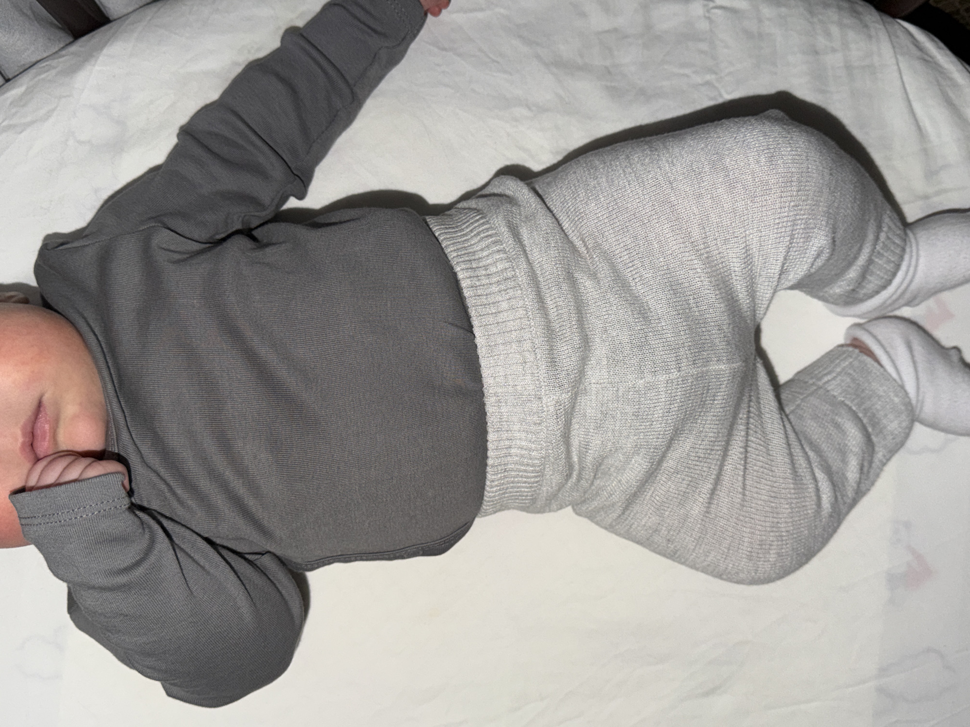 A baby is depicted lying on their back on a white sheet or blanket, seen from a slightly elevated perspective. The baby is dressed in a solid dark grey long-sleeved top and light grey knitted pants with a ribbed waistband. White socks cover the baby's feet. The baby's head is turned slightly to the left, with the face partially visible; the eyes are closed, indicating sleep or rest. One small hand is positioned near the baby's mouth. The overall scene suggests a calm and peaceful moment indoors, possibly in a crib or bed. There are no distinct background elements or visible text. The lighting appears soft and even, typical of an indoor setting. While the location context of "Khimki, Russia" is provided, there are no visual cues within the image itself to confirm this specific geographical location.