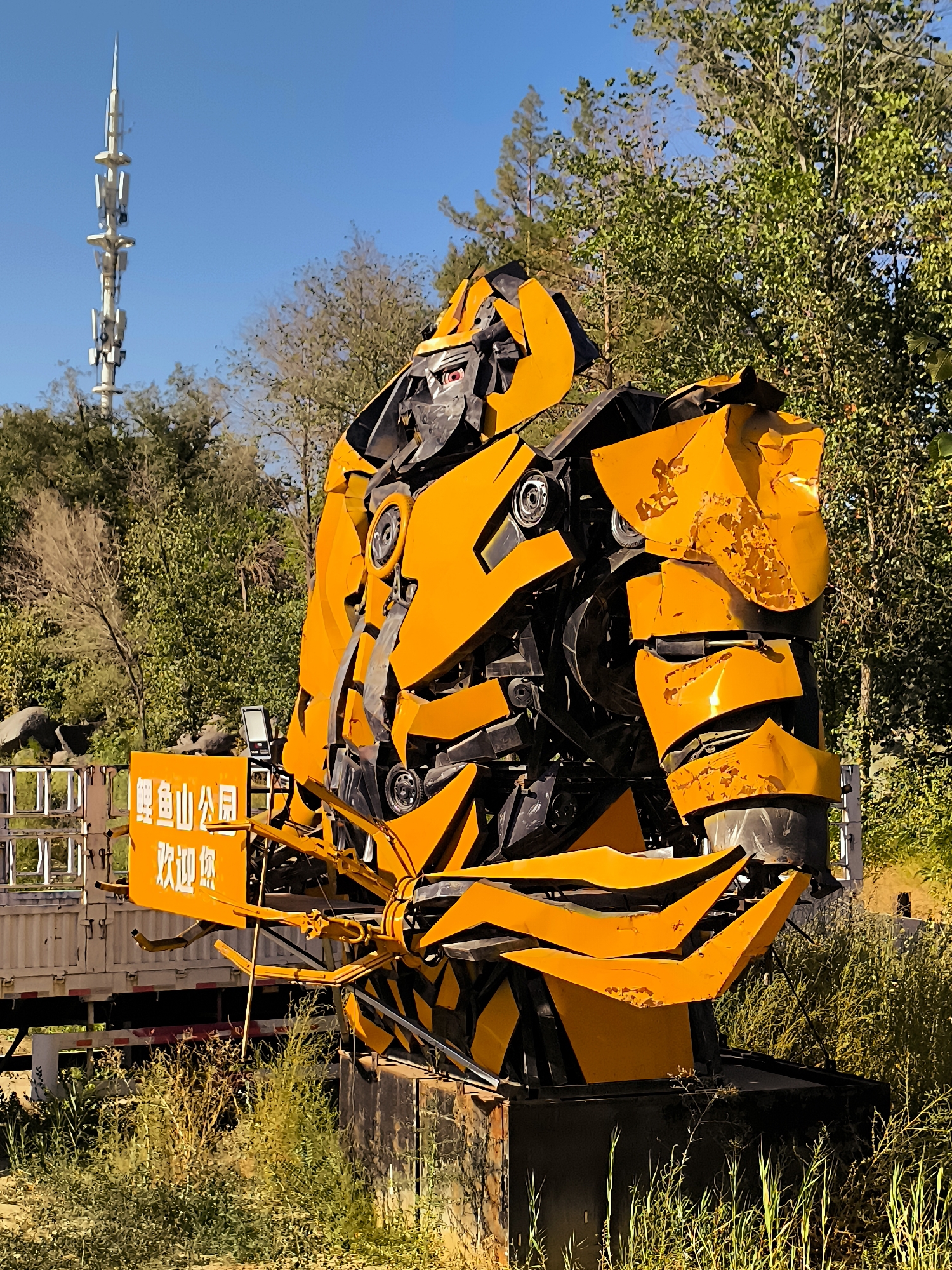 The image depicts a large, bright yellow sculpture resembling a robot, possibly Bumblebee from the Transformers franchise, made from salvaged metal. It stands outdoors in a grassy, overgrown area. In the background, a tall telecommunications tower with multiple antenna arrays pierces a clear blue sky. To the left of the sculpture, a white truck with a flatbed is partially visible, along with a yellow sign bearing Chinese characters that read "鲤鱼山公园 欢迎你" which translates to "Liyushan Park Welcome You." The sculpture itself is composed of angular, distressed metal plates and mechanical components, showing signs of wear and tear. Green foliage and trees surround the sculpture, with some rocks visible in the mid-ground. The lighting suggests it is daytime, with sunlight casting shadows and highlighting the textures of the metal.