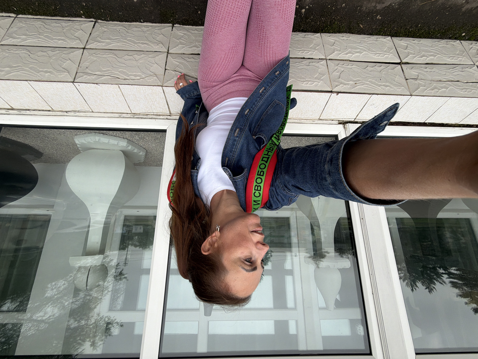 A woman with long, brown hair tied in a ponytail is taking a selfie while sitting on a tiled ledge. She is wearing a denim jacket over a white t-shirt and pink leggings. A green and red strap from a backpack is visible over her shoulder. Behind her are large windows with white frames. Inside the building, white, chess-piece-like sculptures are visible. The reflection in the windows shows trees and a cloudy sky. The tiled ledge is made of textured, light-colored tiles. The ground in front of the ledge is dark and appears to be asphalt or dirt. The scene appears to be outdoors, possibly near a public building. The location is the city of Birobidzhan, Russia.
