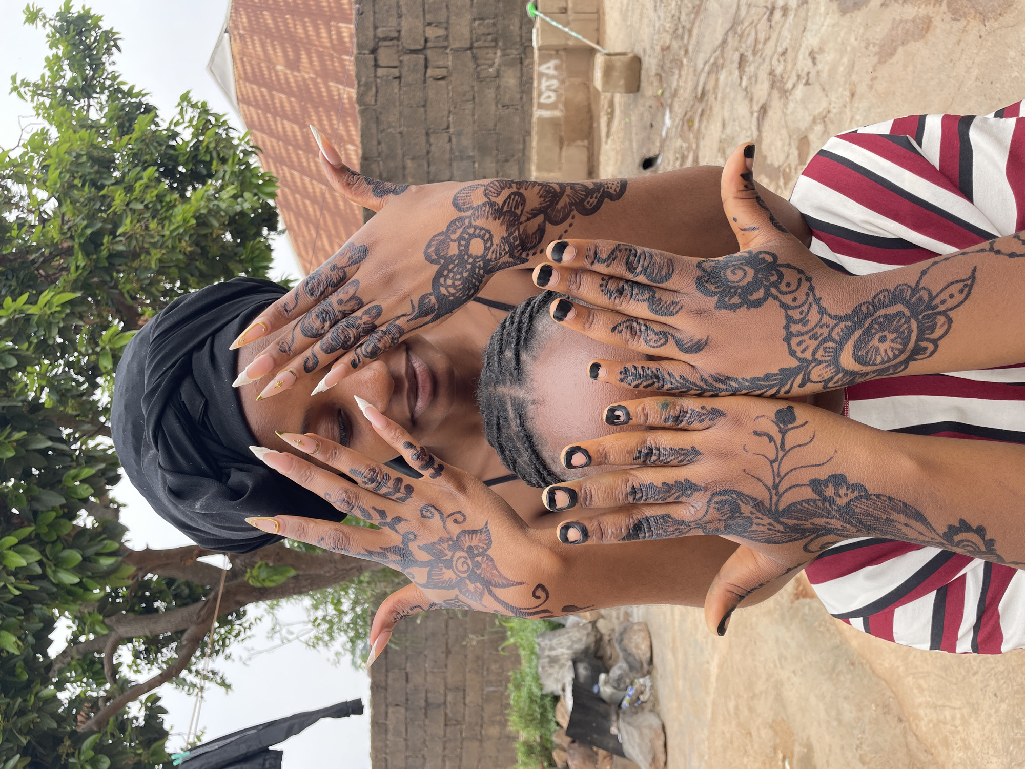 A person with dark skin is centered in the frame, shielding their face with hands adorned with black henna tattoos and nail art. They wear a black head covering. To the right of the person is another individual, positioned lower, whose hands also display henna and black nail polish. The hands of both individuals overlap, partially concealing their faces. The background includes a tree with green foliage, a brick building with a gray hue, and a light-colored ground area. A striped red, white, and black garment is visible in the lower right corner. The location is implied to be near Jos, Nigeria, but specific elements of the city are not discernible in the image.
