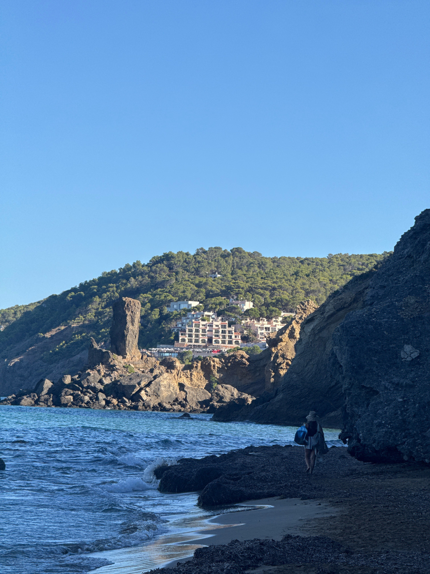 A person walks along a rocky shoreline in Santa Eulària des Riu, Spain. The person is wearing a hat and carrying bags. The water is blue and choppy, with waves breaking on the shore. In the background, there are rocky cliffs covered in green trees, and a building complex is visible on the hillside. The sky is clear and blue.