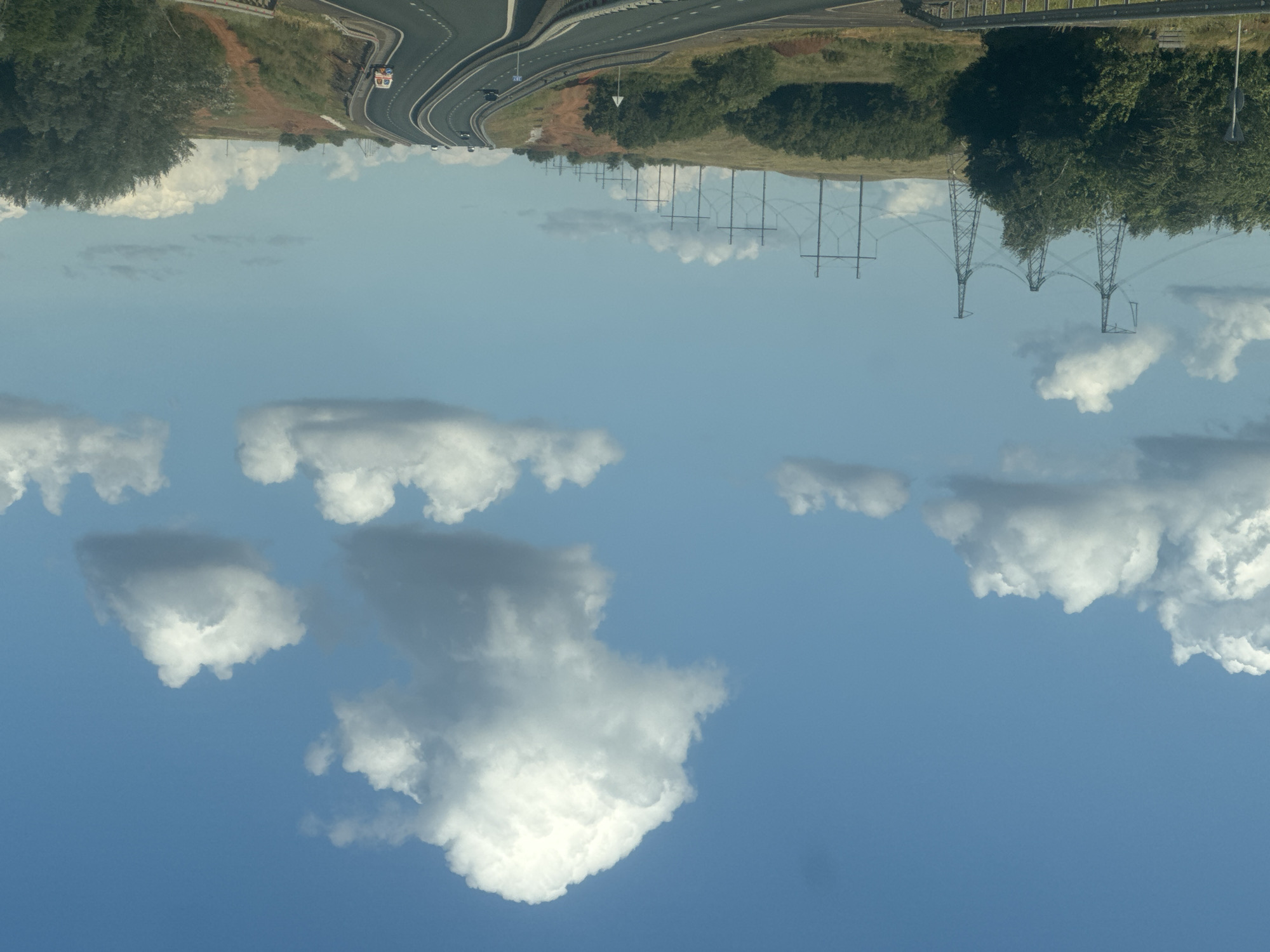 This is a daytime, eye-level, wide-angle shot of a highway in Rodnichnodolsky, Russia. The sky is a clear blue with scattered white cumulus clouds. A paved highway with multiple lanes curves through a hilly, rural landscape. On the left side of the highway, there are dense green trees and tall power line towers. On the right side of the highway, the land is covered in grass and dirt, with more trees in the distance. Several vehicles, including cars and a small bus, are traveling on the highway. The scene depicts a typical road trip through a sparsely populated area with infrastructure for power transmission.