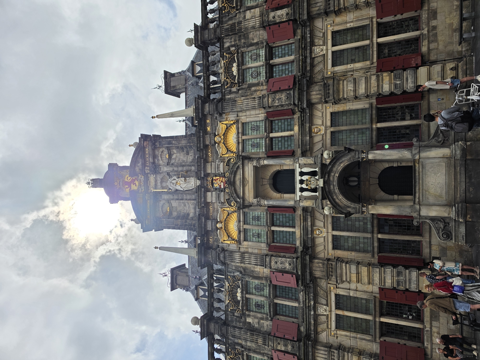 The image shows a wide-angle, low-angle view of the Delft City Hall in Delft, Netherlands. The building is constructed of stone and features ornate architectural details, including gold-colored embellishments, statues, and numerous windows with red shutters. The sun is partially obscured by clouds, casting a bright light on the upper portion of the building.
In the foreground, several people are visible on the sidewalk. On the left, a group of four people are gathered, with two older women sitting on a bench and two younger individuals standing. To the right of this group, a man in a light brown jacket and blue shorts is standing. Further to the right, a man in a blue shirt and dark shorts is looking at his phone, while a woman in a light blue dress and cardigan stands beside him. Near the center-right of the image, a man with a backpack is standing next to a white bicycle, and another man in a light t-shirt and blue shorts is looking at the building.
The sky is cloudy with patches of blue visible, and the sun is a strong, bright light source behind the clouds. The overall scene depicts a bustling urban environment in front of a historical landmark.