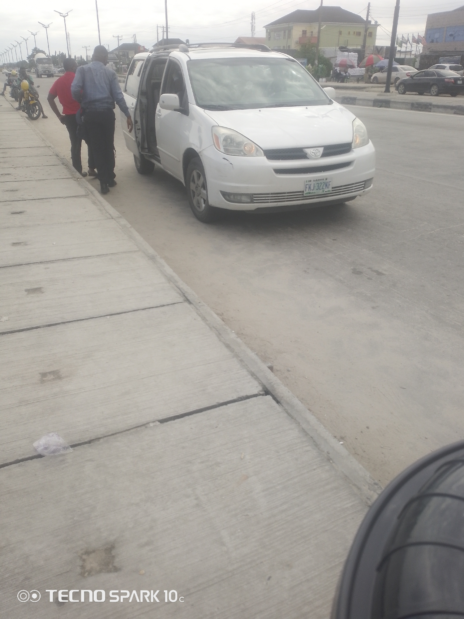 This media file captures a street scene in Lekki, Nigeria, on an overcast day. The primary focus is a white minivan with its side door open, parked on the right side of the road. Two men are standing near the minivan; one, wearing a blue shirt and dark pants, appears to be interacting with the open door, while the other, in a red shirt and dark pants, stands beside him.
In the background, several motorcycles are visible, with one rider wearing a yellow helmet. Further down the road, traffic can be seen, including a truck and other vehicles. Buildings line the background, some appearing to be houses and others possibly commercial establishments. Flags are also visible in the distance. The scene takes place on a paved road with a distinct concrete sidewalk on the left.
The overall atmosphere suggests a mundane moment on a Nigerian street, possibly related to the loading or unloading of the minivan, or a brief stop during travel. There is no indication of specific emotions, but the presence of multiple vehicles and individuals suggests a relatively active urban environment.
Visible text includes a license plate on the minivan reading "FKJ 322KF" and the watermark "TECNO SPARK 10c" at the bottom left of the image. The weather appears to be cloudy, typical for many days in Lagos.
