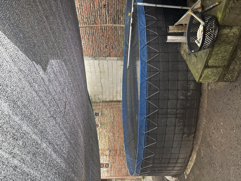 The image displays an outdoor scene featuring a large, round or cylindrical water tank, likely used for aquaculture, positioned between a dark mesh barrier and a series of brick and concrete walls.
On the left side of the frame, a tall, dark gray or black, finely woven mesh or shade cloth extends vertically, covering a significant portion of the view. The texture of this mesh is prominent, with visible individual threads.
To the right of the mesh, occupying the central and right portions of the image, is the large water tank. The tank's main structure appears to be a dark-colored, flexible liner (possibly black geomembrane or thick tarp material) held in place by an external metal grid framework, forming a series of square segments along its side. The top rim of the tank is lined with a vibrant blue tarp material, which is secured to the metal grid with a light-colored rope or wire laced in a zigzag pattern. The tank is partially filled with water, which appears dark. Above the tank, a system of white PVC pipes is visible, with one section featuring a yellow valve, indicating a water inlet or outlet system.
Adjacent to the tank on the right, resting on a stone or concrete block structure, is a black plastic basket with multiple perforations. Inside the basket, several light-colored objects, possibly fish or tools, are visible. Various other miscellaneous items, including what appear to be sections of wooden or PVC pipes, are also arranged on or near this concrete platform.
In the background, behind both the mesh barrier and the tank, there are walls. The lower sections of these walls are constructed from light-colored concrete blocks, while the upper sections are made of exposed red bricks. Some small square or star-shaped openings, possibly vents or decorative elements, are visible within the brickwork. A bamboo pole runs horizontally across the upper part of the brick wall on the far left background.
The ground around the tank and beneath the structures is bare earth or compacted dirt, appearing somewhat uneven. The overall lighting suggests it is daytime, with some shadows indicating the sun's position. The scene depicts a functional, possibly small-scale, outdoor setup for holding water, likely for agricultural or aquacultural purposes.
