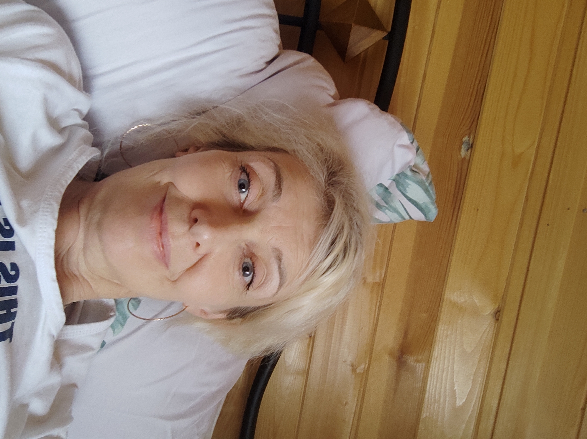 The image shows a selfie of a fair-skinned woman with blonde hair, blue eyes, and fair skin, wearing a white t-shirt. She is lying on a white pillow in what appears to be a bedroom with wooden walls. She is wearing hoop earrings. The background consists of horizontal wooden planks. The overall scene is a close-up of the woman's face and upper body.