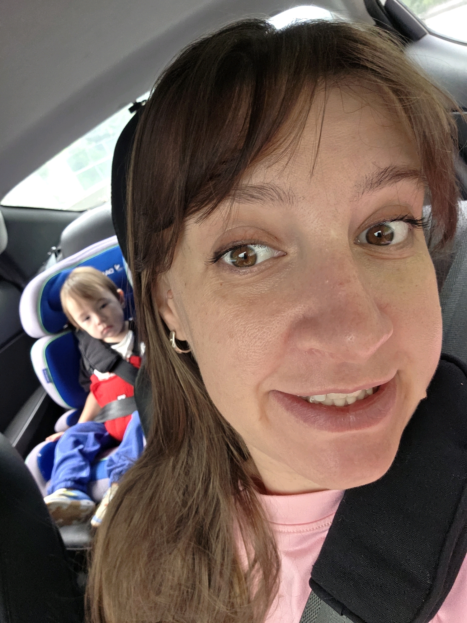 A woman with brown hair and brown eyes takes a selfie in the backseat of a car. She is smiling widely, revealing her teeth. Her left ear is adorned with a small hoop earring. In the background, a toddler sits in a blue and white car seat. The toddler is wearing a red shirt and blue pants. The car's interior is visible, with a gray headrest and a window showing a blurred outdoor scene. The image appears to have been taken during the day.