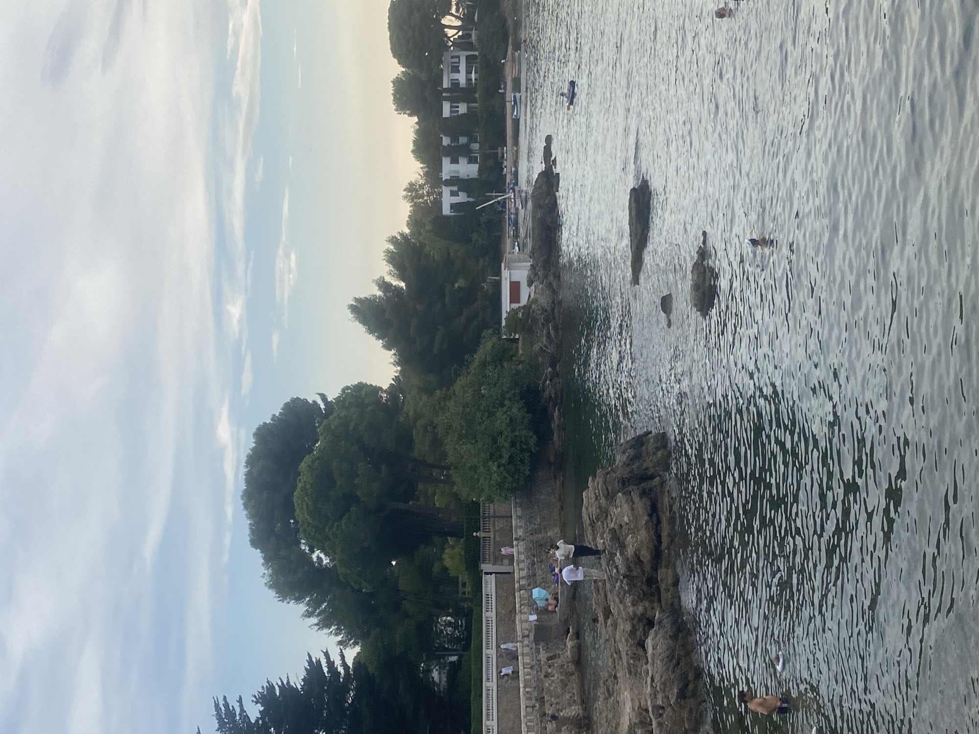 The image depicts a serene waterside scene, likely in Antibes, France, given the architectural style and the Mediterranean vegetation. The foreground is dominated by the calm, rippling water of the sea, with several large rocks scattered in the shallows. On the left, a rocky outcrop juts into the water, where two men are standing and conversing. Further back, a group of people are gathered on a paved area behind a stone wall.
Beyond this area, lush green trees and bushes form a dense backdrop, interspersed with a white building. In the middle ground, a small white building with a red-tiled roof sits close to the water's edge. A few sailboats are moored in the bay, and a white, modern building with multiple windows is visible in the distance, suggesting a residential or hotel complex. Several people are in the water, some swimming and one person in a small boat. The sky above is a soft blue with scattered, wispy clouds, indicating it might be late afternoon or early evening. The overall impression is one of tranquility and leisure by the sea.