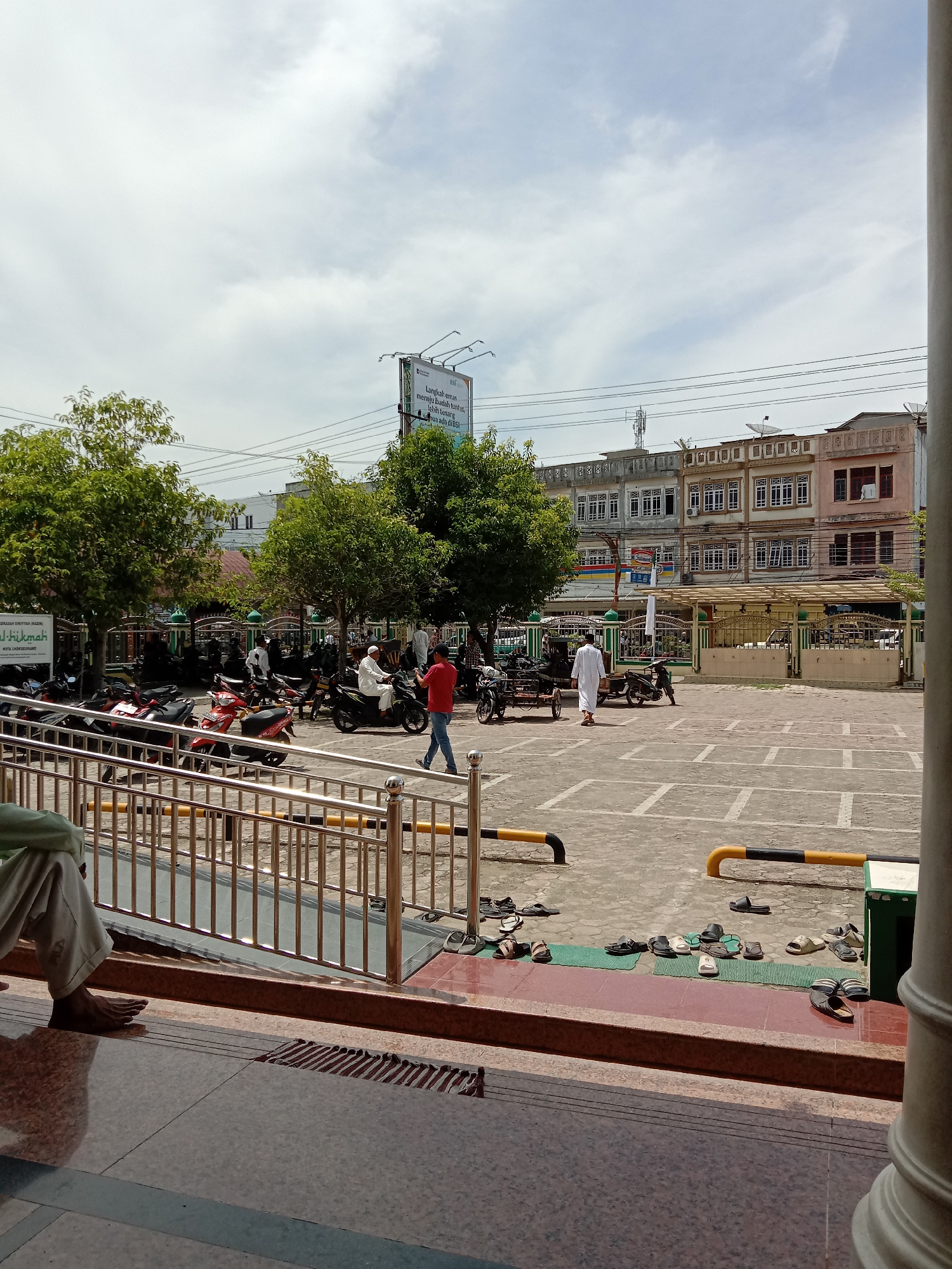 The image shows an outdoor scene in what appears to be a public square in Lhokseumawe, Indonesia.
In the foreground, a metal railing is visible, along with part of a tiled surface, suggesting the edge of a building or structure. There are many pairs of shoes and sandals scattered on the tiled surface and on the ground.
Beyond the railing, a large paved area is visible, marked with parking space lines. Several motorcycles are parked, and a few people are present. One person is walking in a red shirt and blue jeans. Another person is dressed in white clothing, common in some Muslim cultures. Someone on a motorcycle passes.
In the background, trees partially obscure a building with multiple windows, some covered by awnings. A large billboard stands behind the trees. Power lines are strung overhead. The sky is bright with scattered clouds.