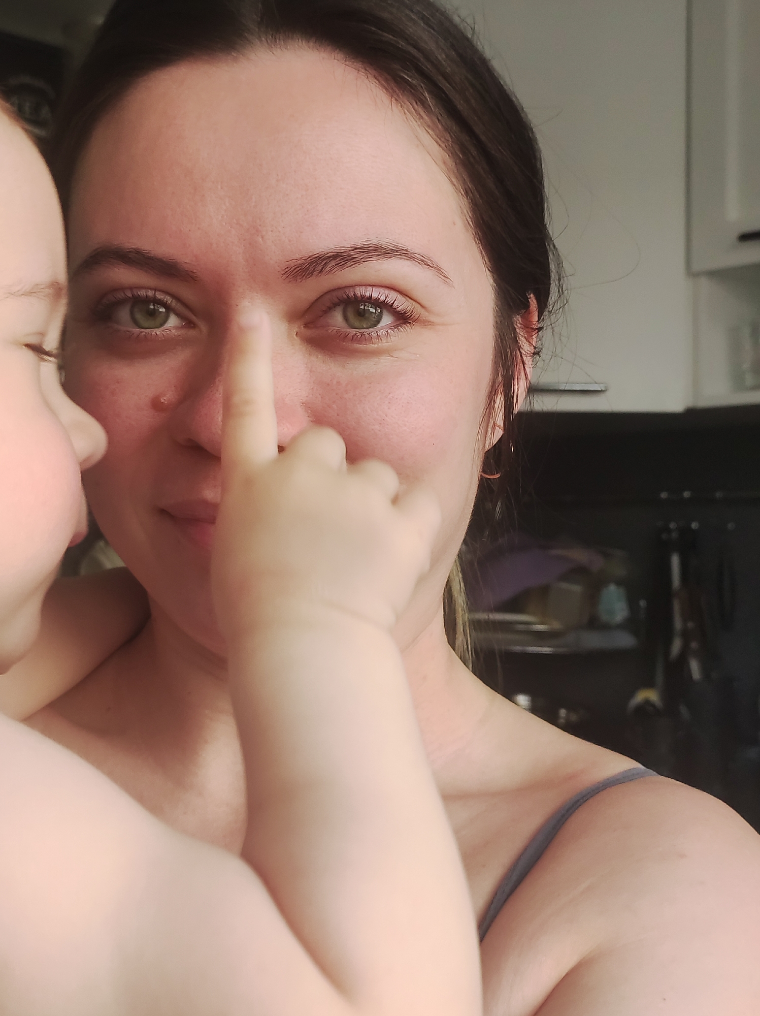 This intimate, close-up photograph, taken in Chaikovsky, Russia, features an adult woman and a baby or toddler engaged in an affectionate interaction indoors. The woman, who has dark brown hair pulled back from her face, smiles warmly while looking directly at the viewer. Her striking green eyes are a focal point, and her skin shows natural texture with a small mole or mark visible on her right cheek/nose area. She is wearing a grey or blue-strapped garment.
On the left side of the frame, the baby's head and arm are visible, with their small hand playfully touching the woman's nose. The baby's skin appears fair and smooth. The lighting suggests it is daytime, with soft natural light illuminating the subjects. In the background, blurred elements consistent with a domestic kitchen setting are visible, including white upper cabinets and darker lower cabinets or shelving units. The scene conveys a sense of warmth, playfulness, and tender connection between the two individuals. No visible text is present in the image.