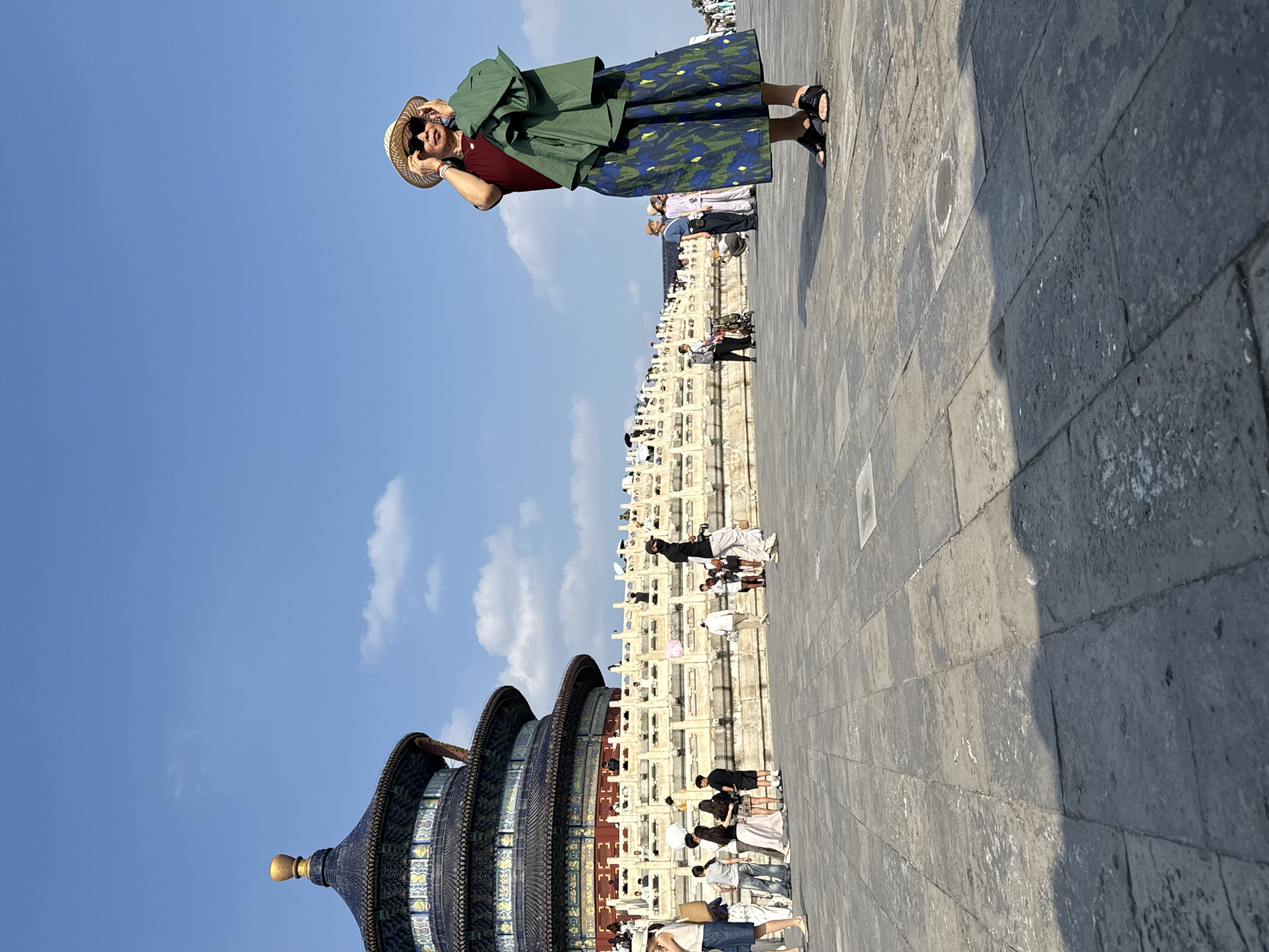 The image shows a woman standing in a large paved courtyard in front of a traditional Chinese temple building. The temple has multiple tiered roofs and is adorned with blue tiles and gold accents. The woman is wearing a straw hat, sunglasses, a dark red shirt, and a long patterned skirt. She is holding a green jacket over her left arm and adjusting her hat. Several other people are visible in the background, walking around the courtyard and near the temple. The ground is paved with large, gray flagstones, and there are shadows cast by the sunlight. The sky is clear blue with a few wispy clouds. This scene is characteristic of the Temple of Heaven in Dongcheng District, China.
