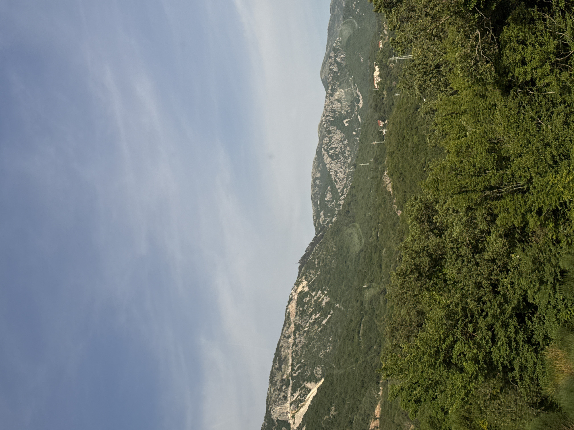 A landscape view shows a mountain range extending vertically. The left portion of the image showcases a blue sky with subtle wisps of clouds. Moving towards the right, the terrain transitions to a series of rocky mountains, interspersed with green vegetation. On the right edge, dense, green foliage predominates. The scene could be located near Markovići, Montenegro. There are no indications of people or human activity, aside from some buildings and power lines visible in the upper-right mountain region.