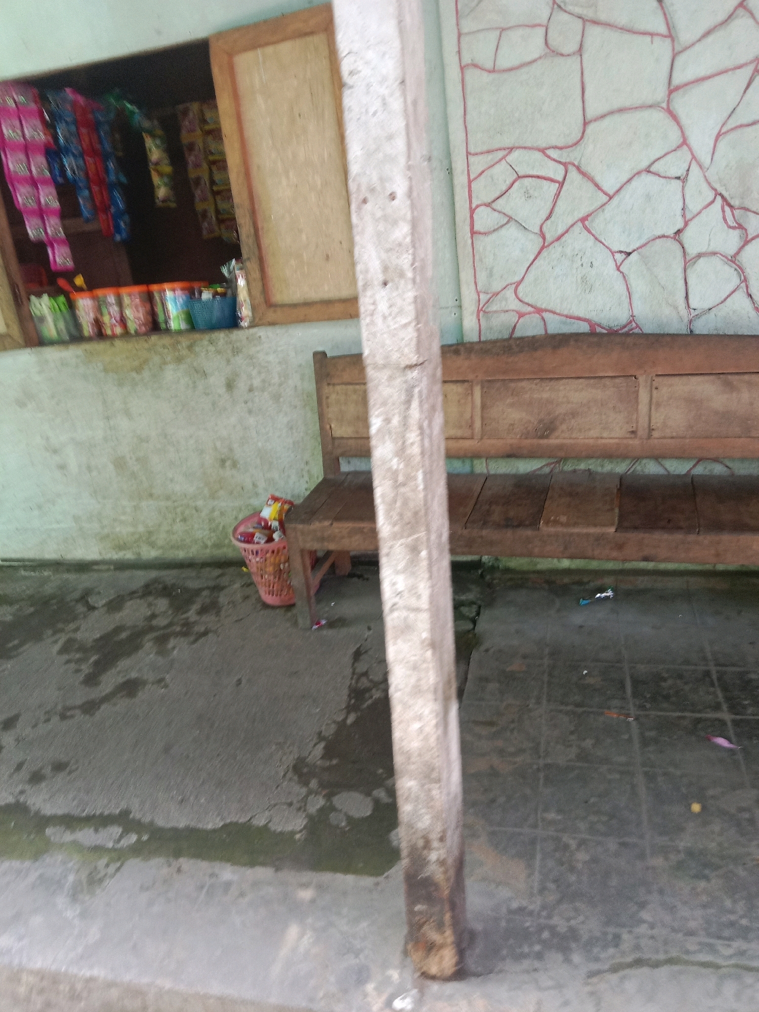 The image depicts an outdoor area in an unknown location in Indonesia, featuring a small storefront with various packaged goods displayed in a window. Adjacent to the storefront, a wooden bench sits on a tiled or paved surface. To the left of the bench and in front of the storefront, a pink basket is filled with what appear to be food wrappers or trash. A concrete pillar stands prominently in the foreground, partially obscuring the view of the bench and the storefront. The wall behind the storefront has a decorative pattern of pink lines on a light-colored background. The ground area appears wet in places, suggesting recent rain or cleaning.