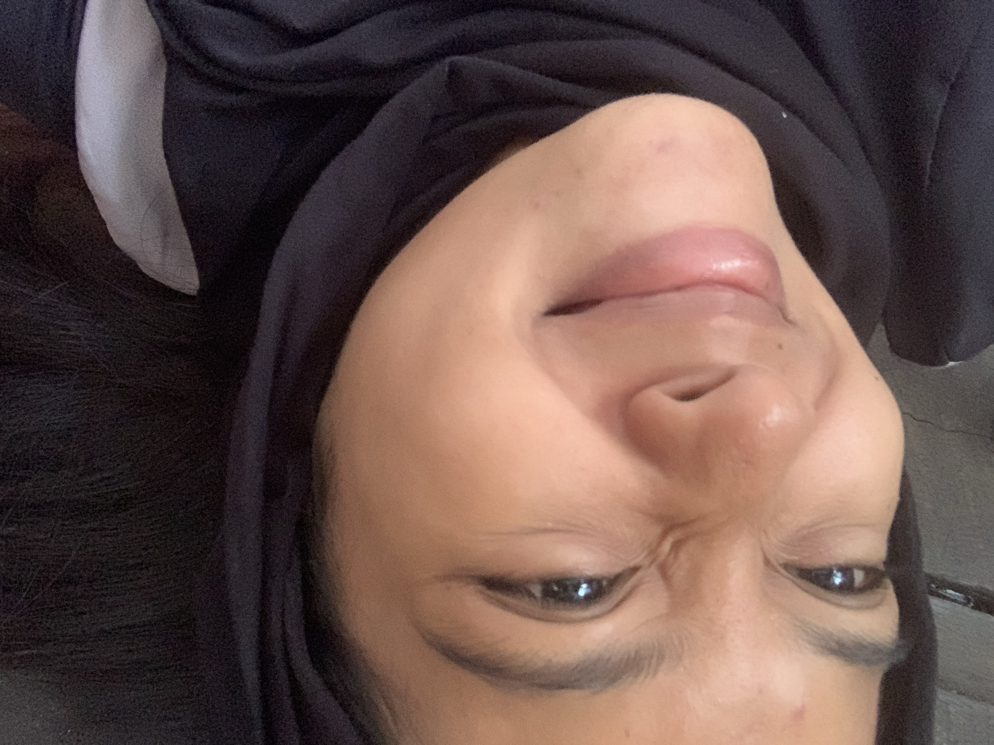 The image is a close-up, upside-down selfie of a young woman wearing a black hijab. She is looking upwards towards the camera with her eyes open and a slightly crinkled nose, giving a playful or perhaps slightly amused expression. Her lips are parted and appear glossy. The lighting is bright, illuminating her skin and highlighting the texture of her face. The background is dark and out of focus, suggesting an indoor or shaded outdoor setting. There are no discernible landmarks or environmental clues within the frame that would indicate a specific location in Sungai Meriam, Indonesia. The focus is entirely on the woman's face and her expression.