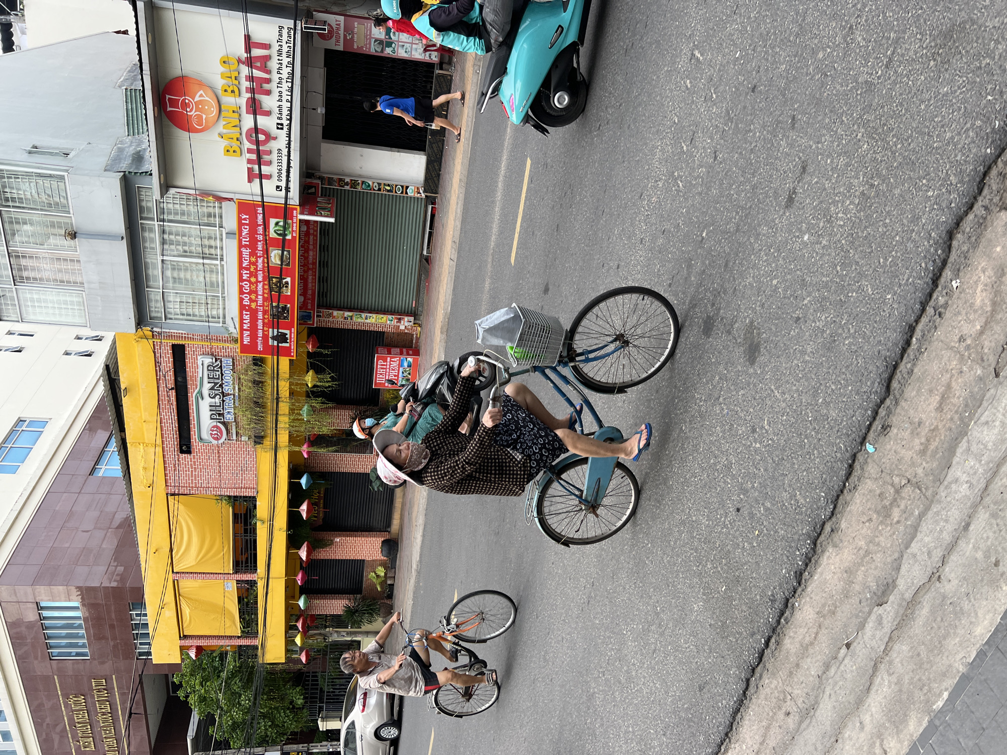 The image captures a street scene in Nha Trang, Vietnam. In the foreground, a woman wearing a patterned long-sleeved shirt, a dark skirt, flip-flops, and a hat is riding a blue bicycle. She has a mask pulled down below her nose. The bicycle has a basket on the front filled with a plastic bag and some green items. A man in a light-colored t-shirt and shorts is riding a bicycle behind her. He is also wearing flip-flops. Further down the street, a person is riding a turquoise scooter. The street is asphalt with a yellow dividing line. Buildings line the street, featuring a yellow facade with red brick accents, awnings, and various signs, including "PILSENER" and "BÁNH BAO THỌ PHÁT". Some of the buildings have balconies and potted plants. Traffic is visible with cars and motorcycles on the road. The sky is overcast.