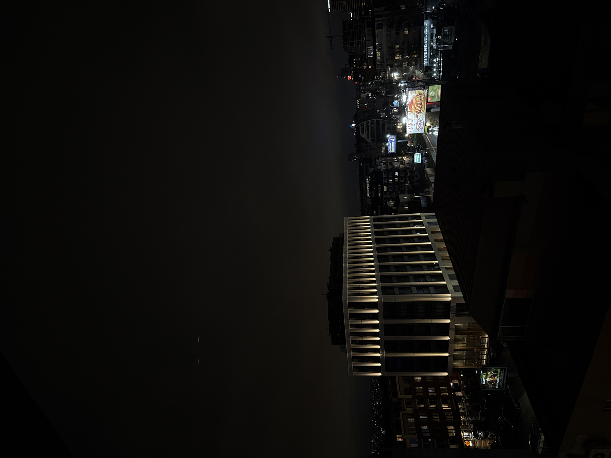 The image depicts a nighttime view of Sofia, Bulgaria. The scene includes multiple buildings illuminated by artificial lights, a city skyline dotted with various structures, and billboard advertisements. The darkness in the sky suggests it's either late evening or night. The image is taken from a high vantage point, looking out over the city.