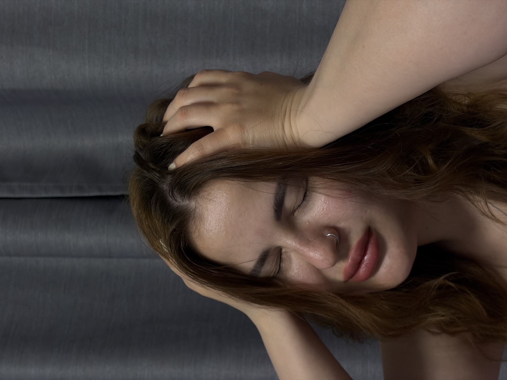 An intimate, tilted close-up shot captures a young woman, likely in Krasnodar, Russia, with her eyes closed, suggesting a moment of intense emotion, contemplation, or sensual pleasure. Her head is tilted, and her long, wavy brown to auburn hair frames her face. Her lips are slightly parted, revealing a subtle smirk or relaxed expression, enhanced by what appears to be lip gloss or a natural sheen. She has a small silver hoop nose ring on her right nostril.
Her hands are prominent in the composition, with fingers intertwined in her hair, seemingly grasping or running through it, adding to the emotional depth of the scene. Her skin appears fair, and a hint of makeup is visible, particularly on her eyebrows. The nails on her visible fingers are painted a light color, possibly white or pale pink. The background is a plain, dark grey textured fabric, possibly a curtain or bedding, which provides a soft, uncluttered setting and allows the focus to remain entirely on the woman. There is no visible text in the image. The lighting appears soft and indirect, contributing to the private and intimate atmosphere.