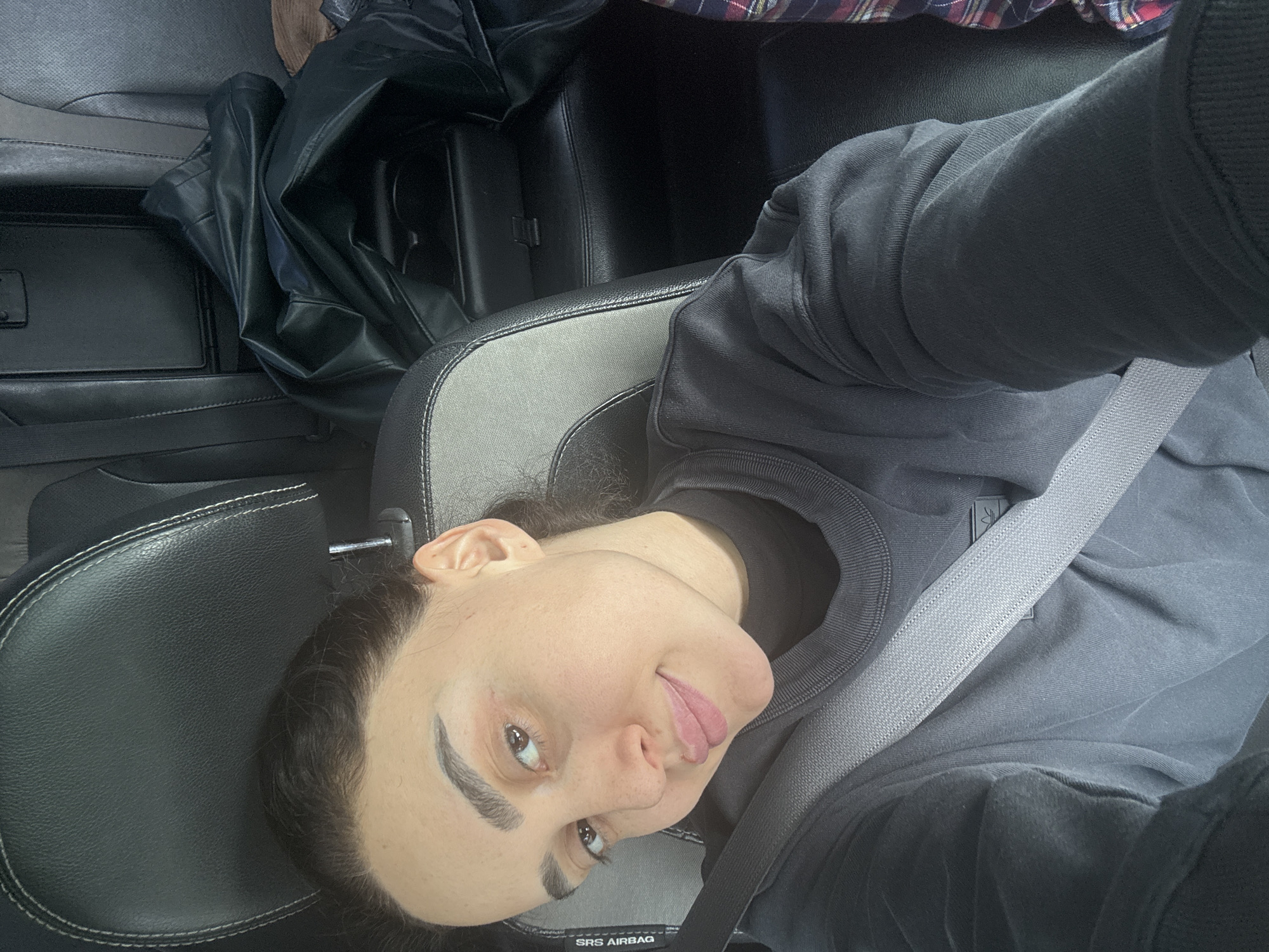 A person with dark hair and light skin is in a car, wearing a grey sweatshirt and a seatbelt. The car has black leather seats with grey accents. A black leather jacket is in the back seat. The location is likely Tbilisi, Georgia, although there is no definitive evidence in the image to confirm this.