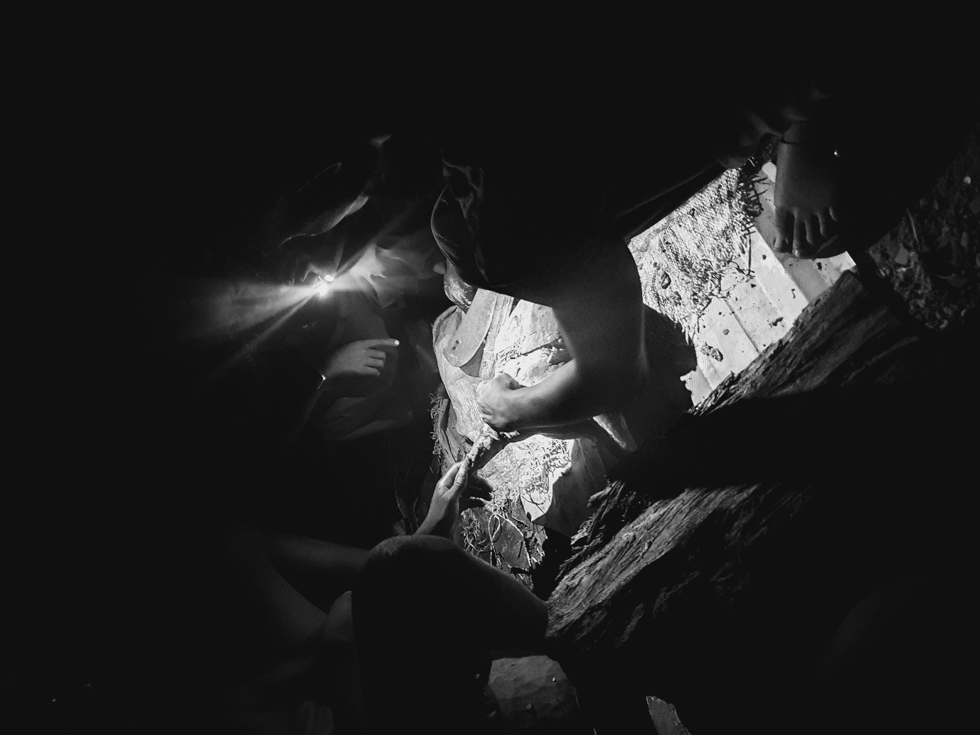 A black and white, low-angle shot depicts a group of people congregated around a light source in what appears to be an underground space, possibly a cave or a tunnel. A person holding a light directs the beam towards a central point where others are gathered. The scene is dimly lit, with surrounding areas shrouded in darkness. Hands and faces are partially visible, suggesting activity or interaction involving objects in the lighted area. The presence of exposed feet indicates some individuals may be barefoot. Debris or rough-hewn materials, possibly rocks or wood, surround the perimeter of the light. There is no identifiable reference to Idah, Nigeria.