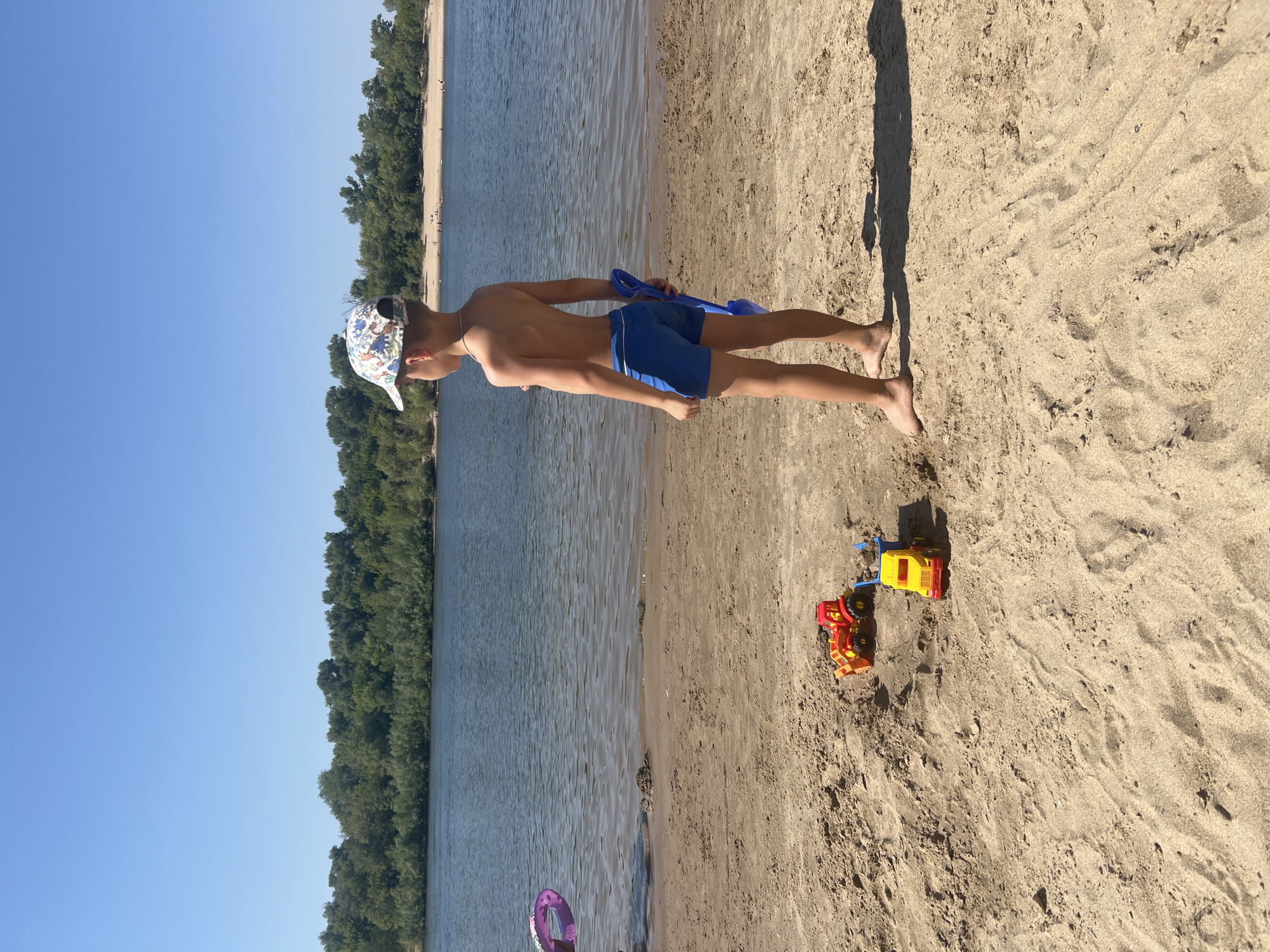 In Kilyakovka, Russia, on a sunny day, a young boy stands on a sandy beach next to a body of water. He is wearing blue swim trunks and a patterned cap, and is holding a blue shovel. In front of him, two toy construction vehicles, a red and yellow bulldozer and a yellow and blue dump truck, are partially buried in the sand. The water is calm, and across the river, a dense line of trees forms the treeline. The sky is clear and blue.