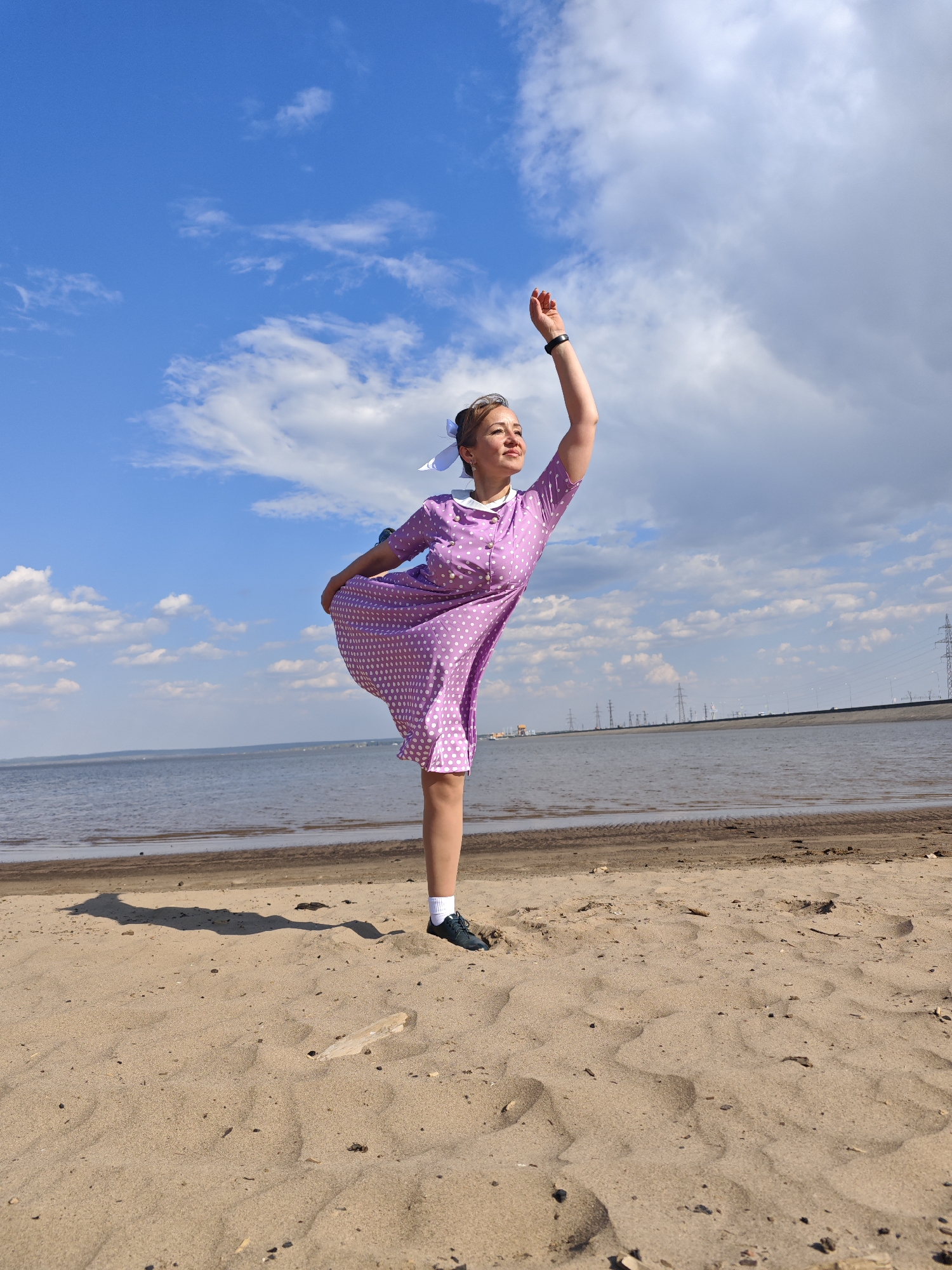 A woman stands in a dancer's pose on a sandy beach near the Gulf of Finland in Noviy, Russia. She is wearing a lavender polka-dot dress with a white collar and bows in her hair. Her right arm is extended upwards, and her left hand holds the side of her dress, lifting it slightly. The beach slopes down towards the water. In the background, the Gulf of Finland stretches to the horizon, with a low-lying coastline visible in the distance. Electrical towers can also be seen. The sky is blue with scattered clouds.