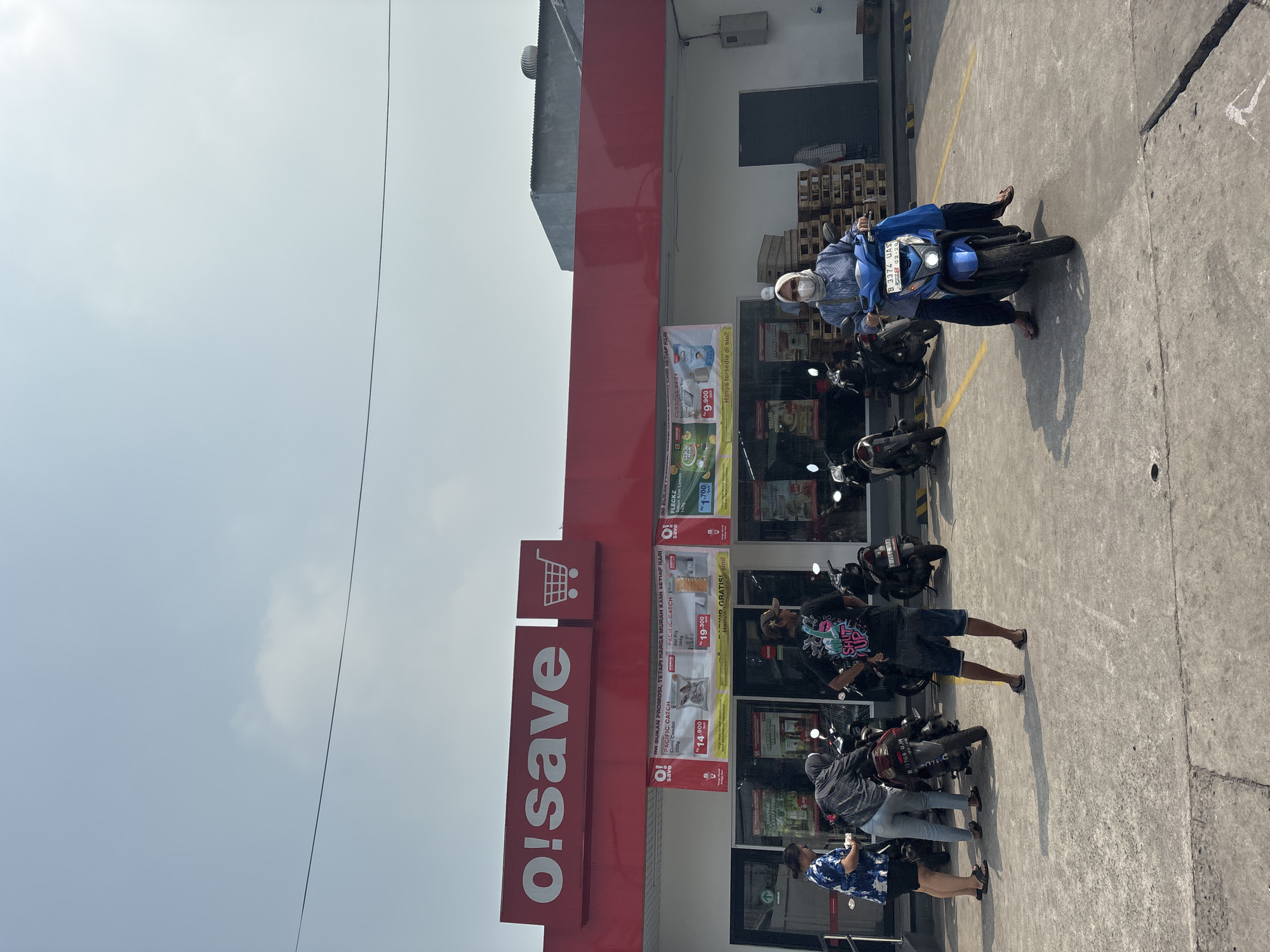 In North Jakarta, Indonesia, O!save supermarket's red facade is visible under a pale blue sky with a thin, dark line stretching horizontally across it. Several people are gathered in the paved outdoor area in front of the store, some standing near motorcycles and others interacting. A woman in a blue and white patterned shirt and dark shorts stands on the left, facing away from the camera, examining something in her hands. Beside her, a person in a dark grey hoodie and light blue pants also looks down. A man wearing a black t-shirt with "SHUT UP" printed on it in pink and white, denim shorts, and a brown baseball cap stands with his back mostly to the camera, his right hand on his hip. He is positioned between two parked motorcycles. In the foreground, a woman wearing a light blue hijab, glasses, and a matching blue long-sleeved shirt with dark pants rides a blue motorcycle, facing towards the right side of the frame. Her face is partially obscured by a light-colored mask. The motorcycle has a blue license plate with white text that reads "B 3374 UAS". The ground is concrete with yellow painted lines marking parking spaces. Pallets are stacked to the right of the store entrance, and a large ventilation unit is visible on the roof of the building in the background. The entrance to the store features glass doors and several promotional posters displaying product prices.