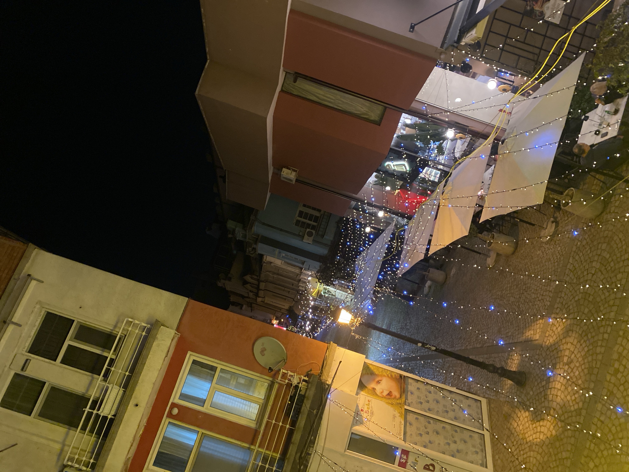 The image shows a street scene in Plovdiv, Bulgaria, at night. The camera is tilted at an unusual angle. Buildings line the street, with windows and architectural details visible. A lamppost is present, casting light onto the cobblestone pavement. Above the street, strings of blue and white lights are strung across, and white fabric sail-like decorations are suspended. There is some signage and advertising visible on the buildings. The sky is dark.