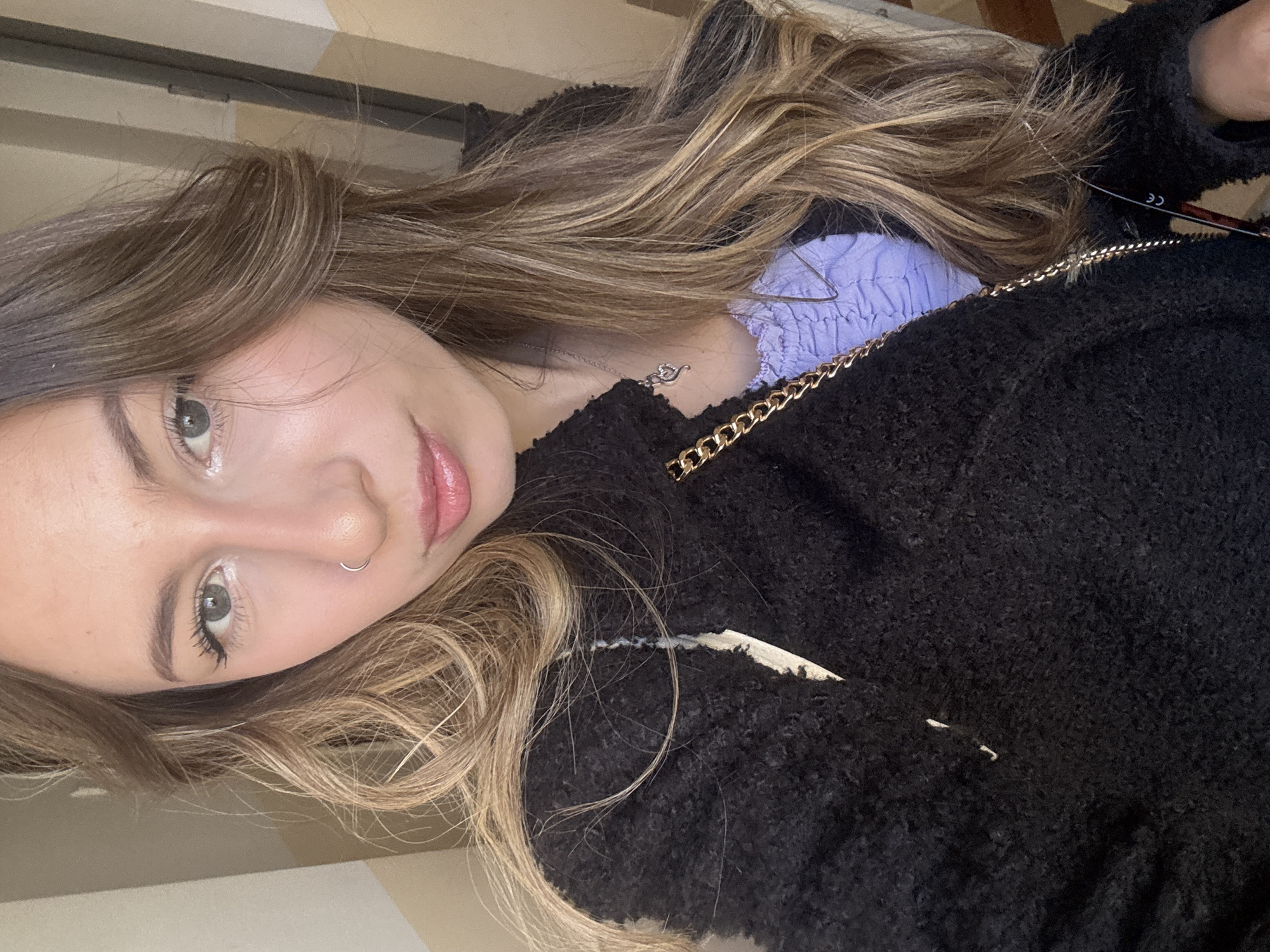 A close-up portrait shows a woman in an indoor setting, with the image captured from a tilted perspective. She has long, wavy hair that is predominantly brown with distinct blonde highlights, framing her face. Her light-colored eyes, appearing green or hazel, gaze directly towards the viewer, enhanced by visible eyelashes. She wears light makeup, including a pink lip gloss, and a small silver hoop nose ring on her left nostril.
She is dressed in a black, textured, and possibly fuzzy jacket, layered over a light purple top with ruched detailing on the neckline. A delicate silver-colored necklace with a small pendant, resembling a heart or infinity symbol, rests on her neck. A prominent gold-colored chain, likely an accent on her jacket or a strap from a bag, runs across the upper part of her torso.
The background consists of light-colored walls or a ceiling, suggesting an interior location. The lighting appears to be natural, possibly daylight, illuminating her face. A small white tag with the letters "CE" is partially visible on the black jacket near the shoulder. The photograph is taken in Veliko Tarnovo, Bulgaria.