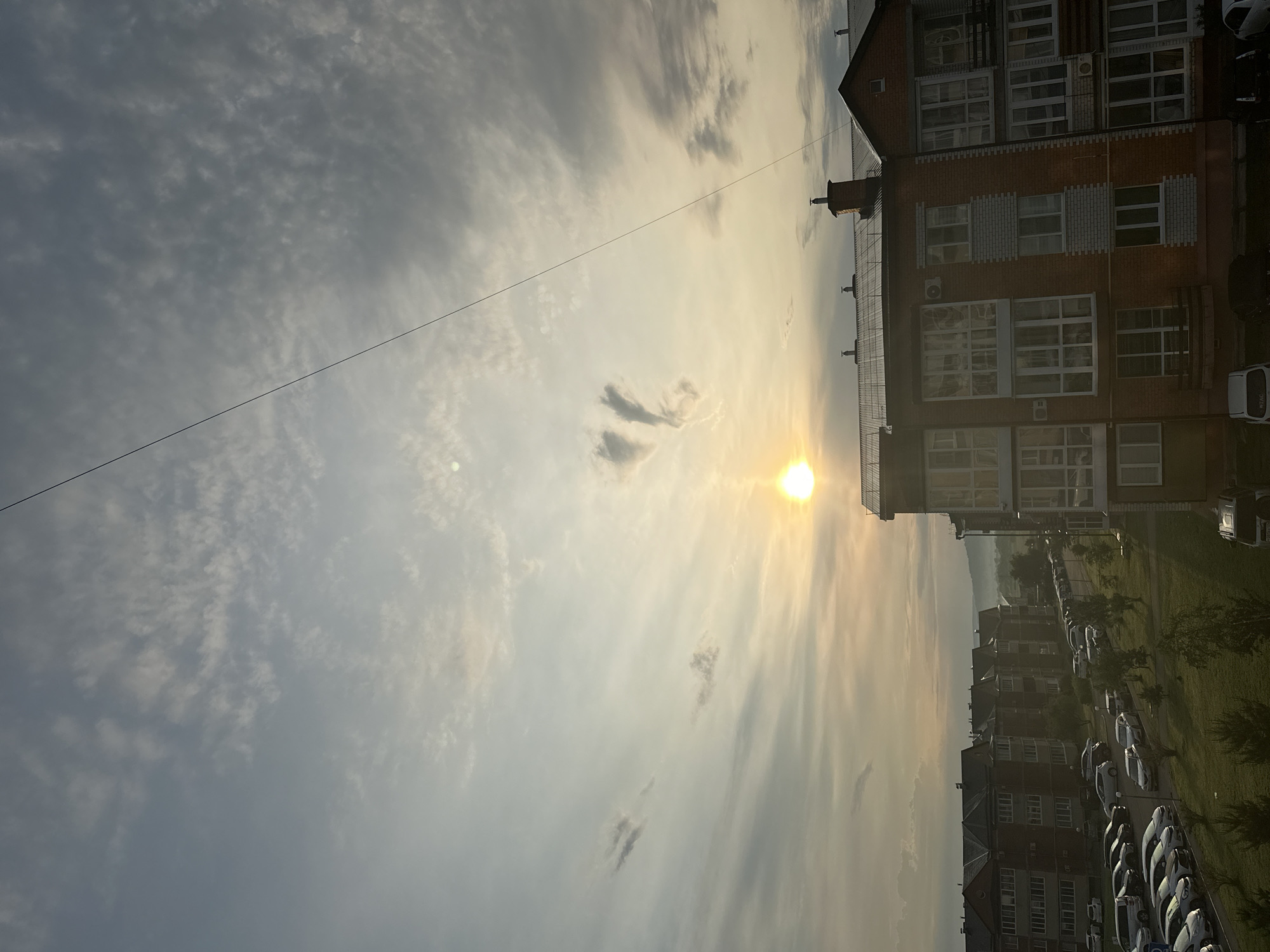 This is a high-angle, medium shot of a residential area in Ussurijsk, Russia, during a hazy sunset. The sky is filled with scattered, thin clouds, and the sun, a bright orb, is positioned in the upper left quadrant. A thin black wire stretches diagonally across the frame.
In the foreground and midground, several multi-story brick apartment buildings are visible. These buildings have numerous windows, some of which are reflecting the light from the sky. Balconies are also present on some of the buildings. The architecture suggests a modern, urban environment.
Below the apartment buildings, a street runs through the scene, lined with parked cars. The cars are mostly sedans and SUVs, in various colors. Green grass and trees are interspersed throughout the area, indicating a presence of vegetation within the city. In the background, rolling hills or low mountains can be seen, partially obscured by haze.
There are no people directly visible in the image. The scene captures a quiet, everyday moment in a Russian city, with the soft light of sunset creating a diffused and slightly ethereal atmosphere. The presence of parked cars and apartment buildings clearly identifies it as an urban or suburban residential setting.