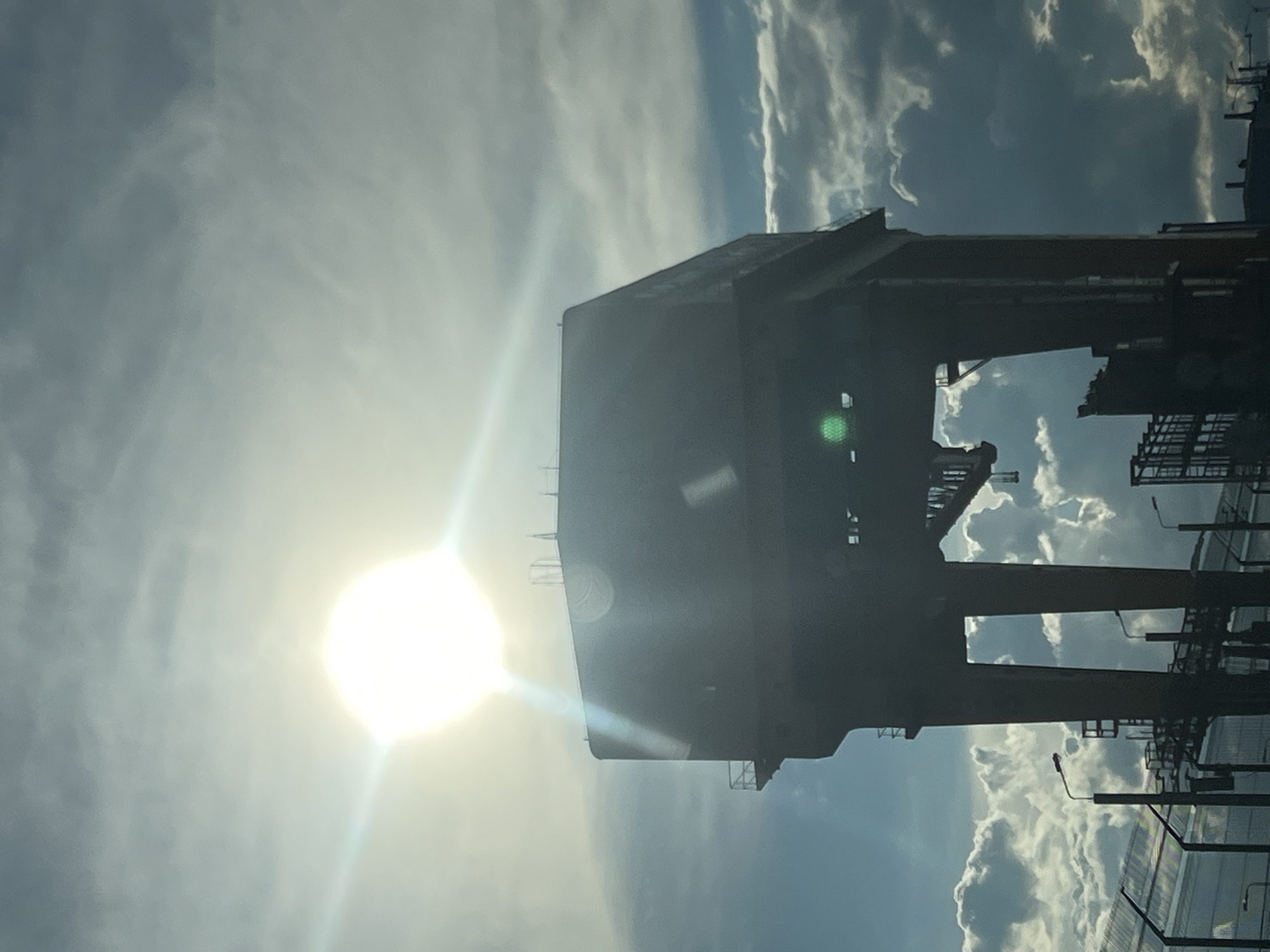 The image shows a large gantry crane silhouetted against a bright sun and cloudy sky. The crane is complex in structure with several interconnected platforms and supporting beams. Adjacent to the crane are street lights and fencing. This image was taken in Chaikovsky, Russia.