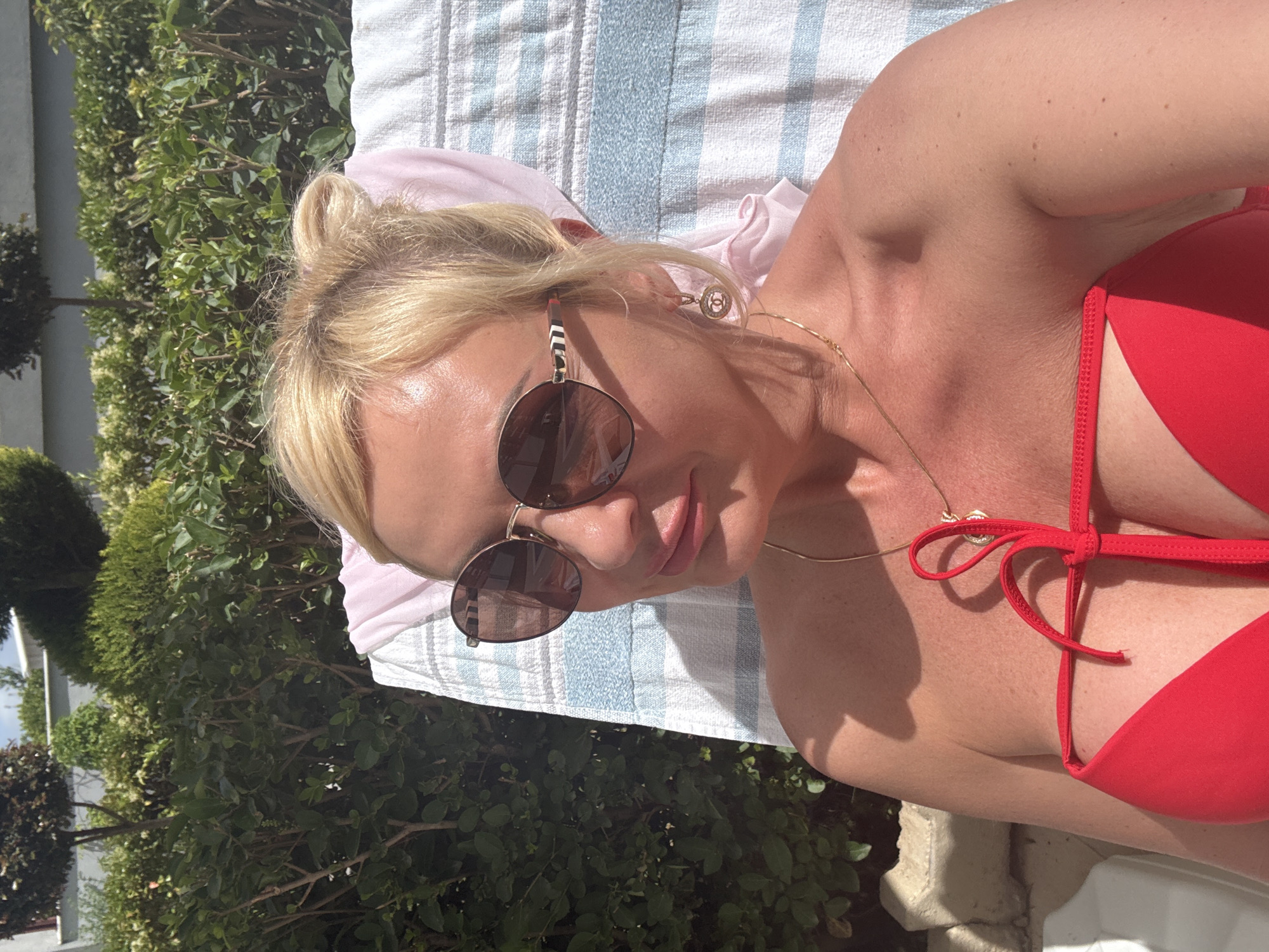 A woman lies on a striped towel in an outdoor setting. She is wearing a red bikini and sunglasses, with her blonde hair pulled up in a bun. A gold necklace with a charm is visible on her chest. The area surrounding her features green bushes. The location appears to be in a sunny, outdoor area, possibly in the city of Unknown, Unknown.